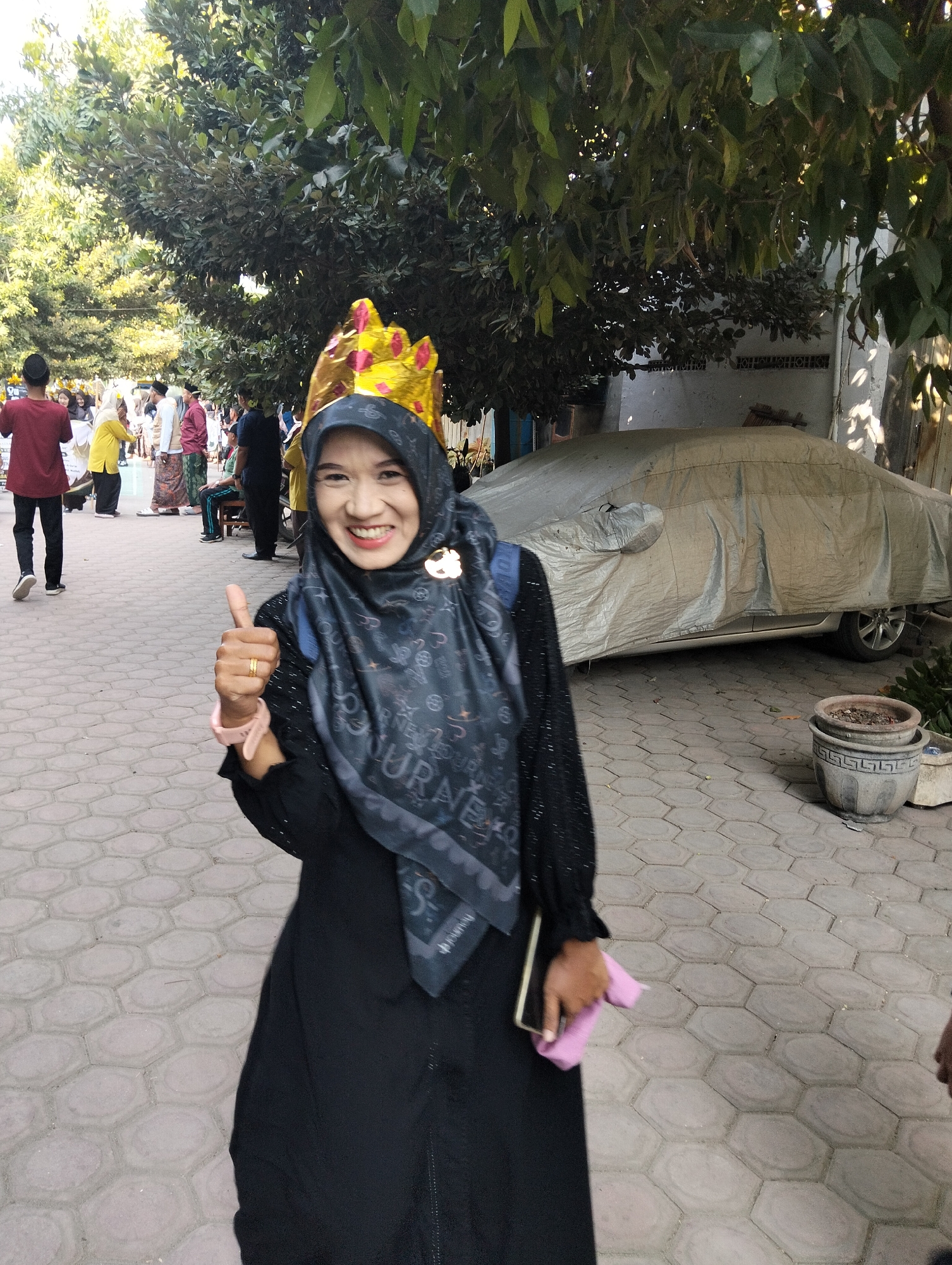 A woman wearing a black hijab and a golden crown with red accents stands on a hexagonal paved walkway in Kranji, Indonesia. She is smiling and giving a thumbs-up with her right hand, which has a gold ring on her ring finger and a pink watch on her wrist. She is holding a pink cloth and a smartphone in her left hand. She is wearing a black abaya with black sequins on the sleeves. In the background, a car covered with a light-colored tarp is parked. Trees with green leaves are visible in the upper portion of the image. Several other people are walking and standing in the background. The sun is shining, casting shadows on the ground.