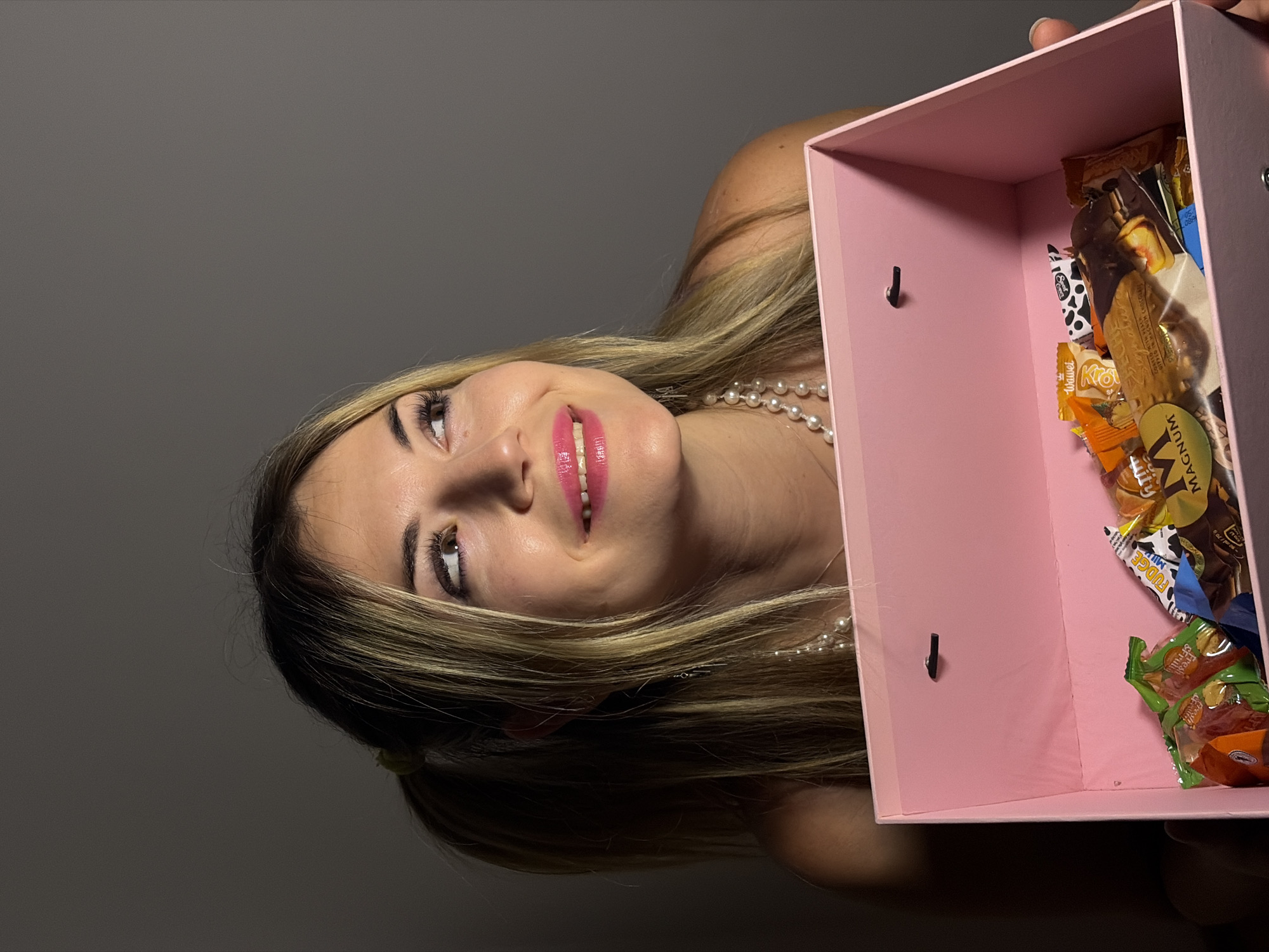 A woman is holding a pink box full of candy. The woman has blonde hair and is wearing a pearl necklace. The box is open, and the candy is visible inside. The background is a neutral grey color. There is no indication this image was taken in Budva, Montenegro.