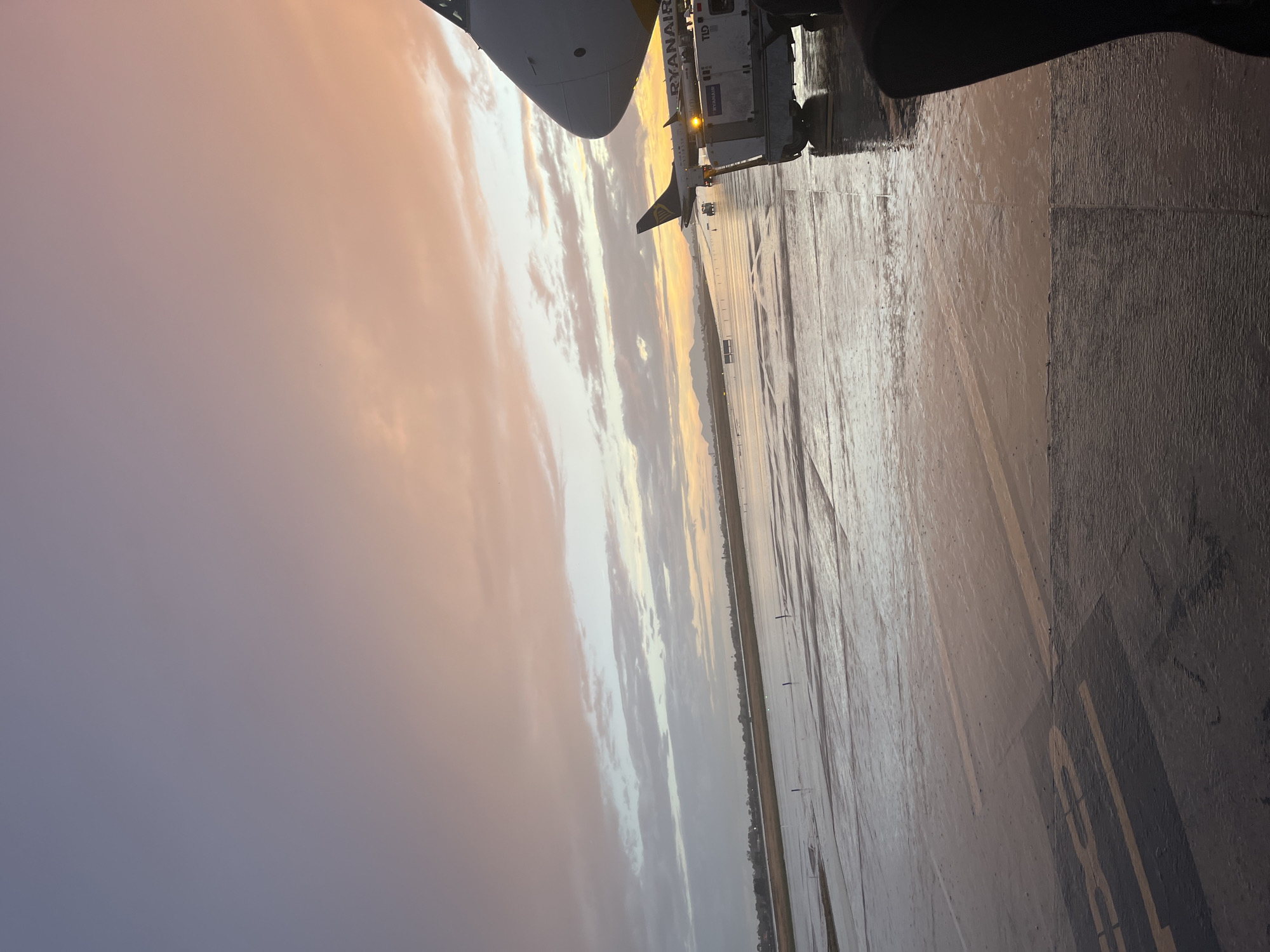 The image shows a partial view of an airplane, specifically a Ryanair plane, at what appears to be an airport apron. The ground is wet, possibly from rain, and there are markings on the pavement indicating taxiways or parking areas. In the background, there is a gradient sky indicating either dawn or dusk, with pastel colors blending from purple to orange. A ground service vehicle, possibly used for baggage loading or other support operations, is connected to the airplane. The location is likely Alicante-Elche Airport (ALC) given Ryanair's presence.