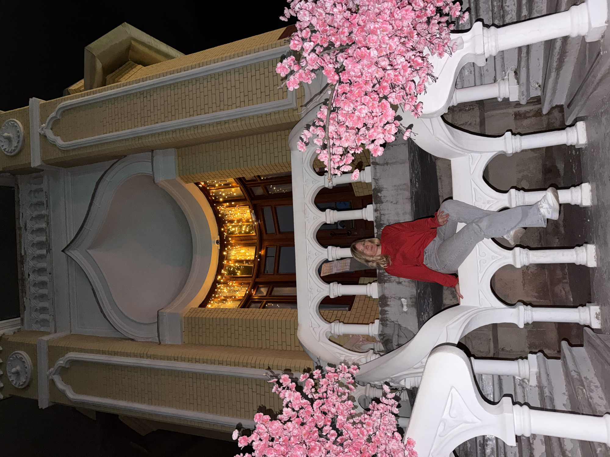A woman with blonde hair, wearing a red long-sleeved shirt and grey pants, sits on a white ornate railing in front of a building. The building is made of light yellow bricks and has decorative white trim and arches. String lights are visible behind a large window. Two artificial cherry blossom trees with pink flowers flank the staircase. The scene is set at night, with the building illuminated. This building is the Narzan Gallery in Kislovodsk, Russia.