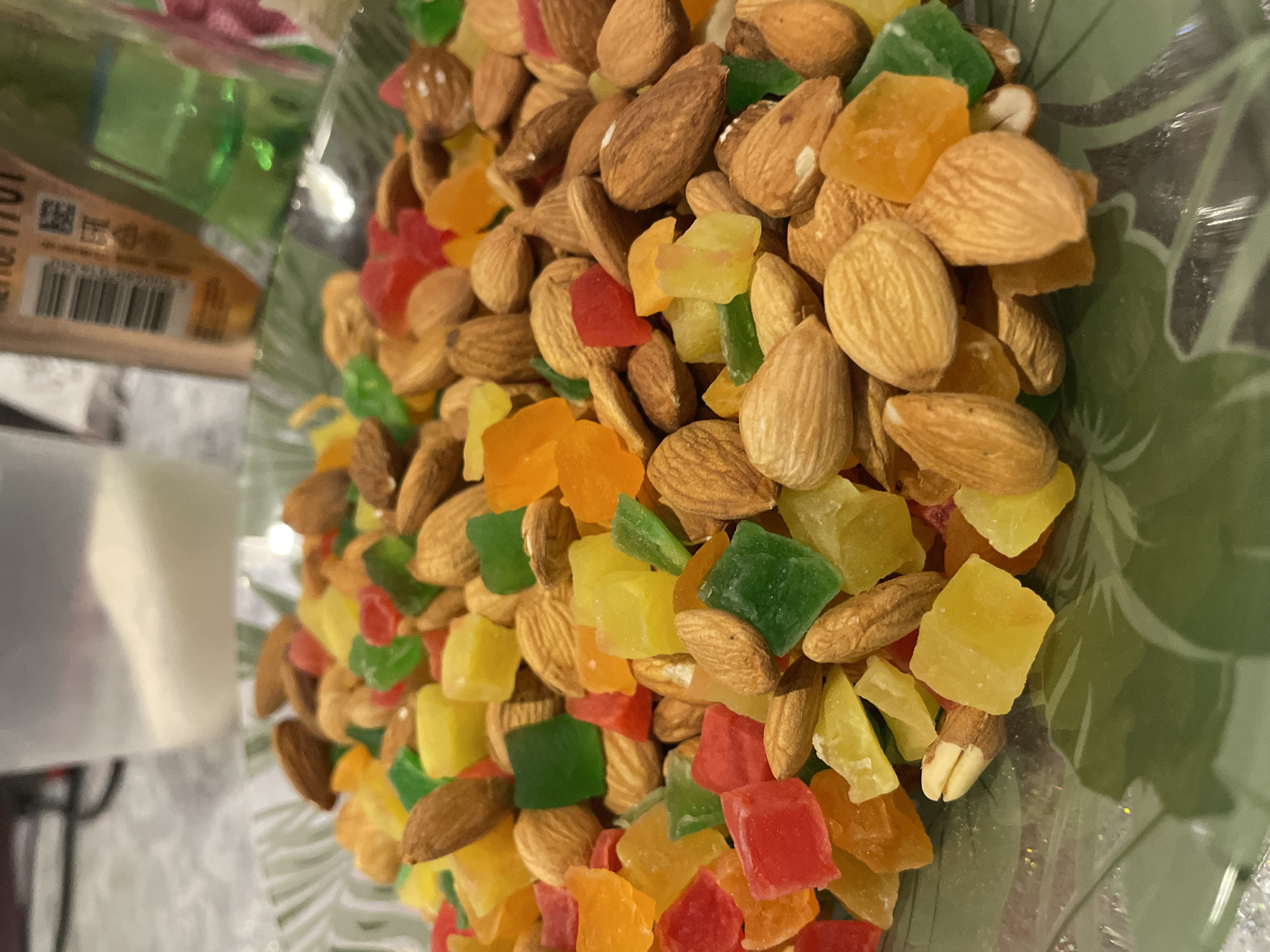 A close-up, slightly angled, high-resolution image displays a heap of almonds mixed with diced candied fruit. The almonds are mostly whole, with brown skins. The candied fruit pieces are brightly colored, including red, green, orange, and yellow hues. The almonds and candies are in a glass bowl with a green leaf design. The bowl sits on a surface near a package labeled "Settle." The location is not discernible from the image, and therefore cannot be pinpointed to Olginka, Russia.