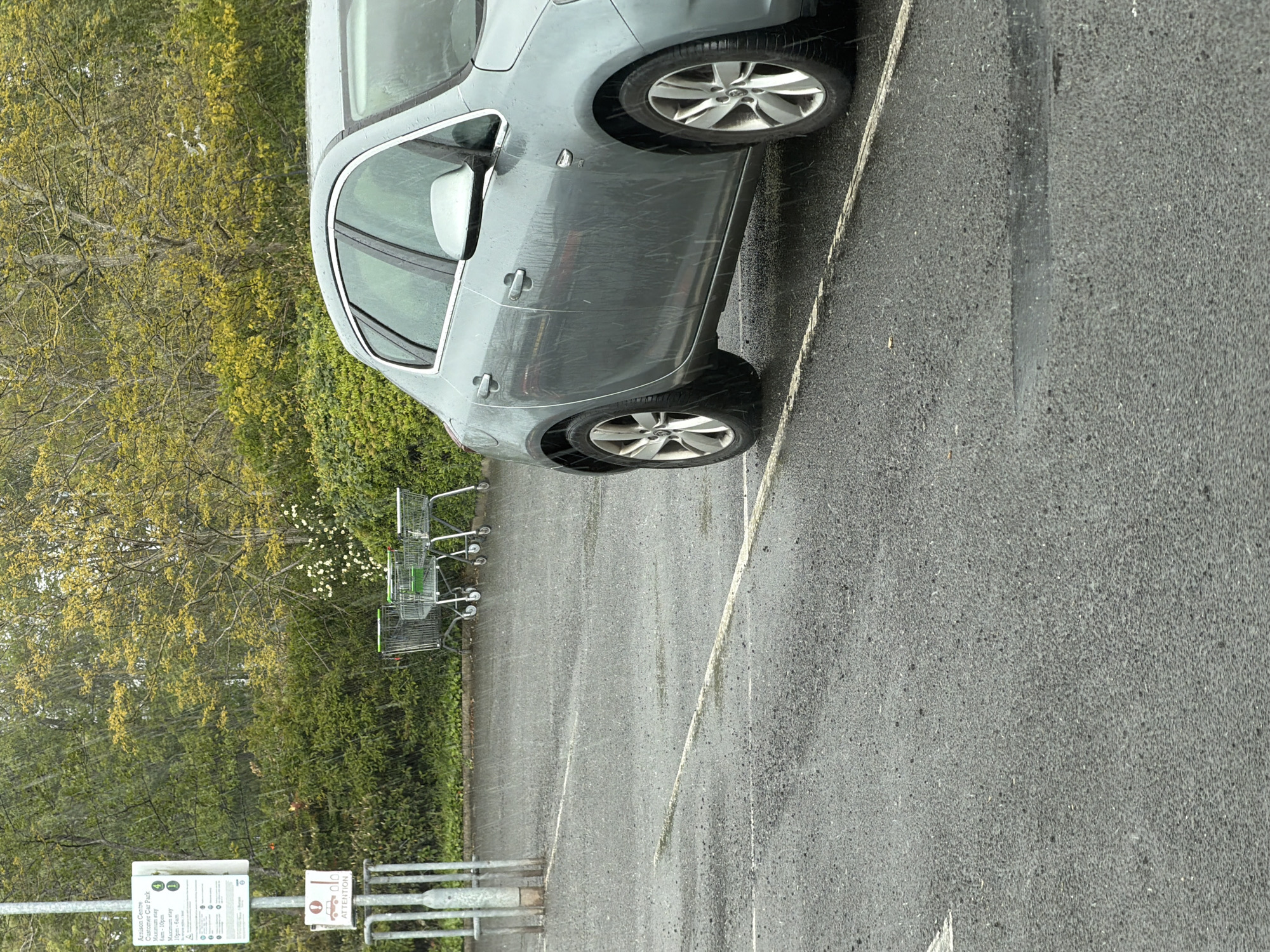 The image shows a gray car parked in a parking lot in Pity Me, United Kingdom. The parking lot is lined with a row of trees and bushes. There are several shopping carts stacked next to the parking lot. There is a sign that reads "Arnison Centre Customer Car Park Maximum stay 6am-10pm Maximum stay 10pm-6am." The parking lot appears wet, and it may be raining.