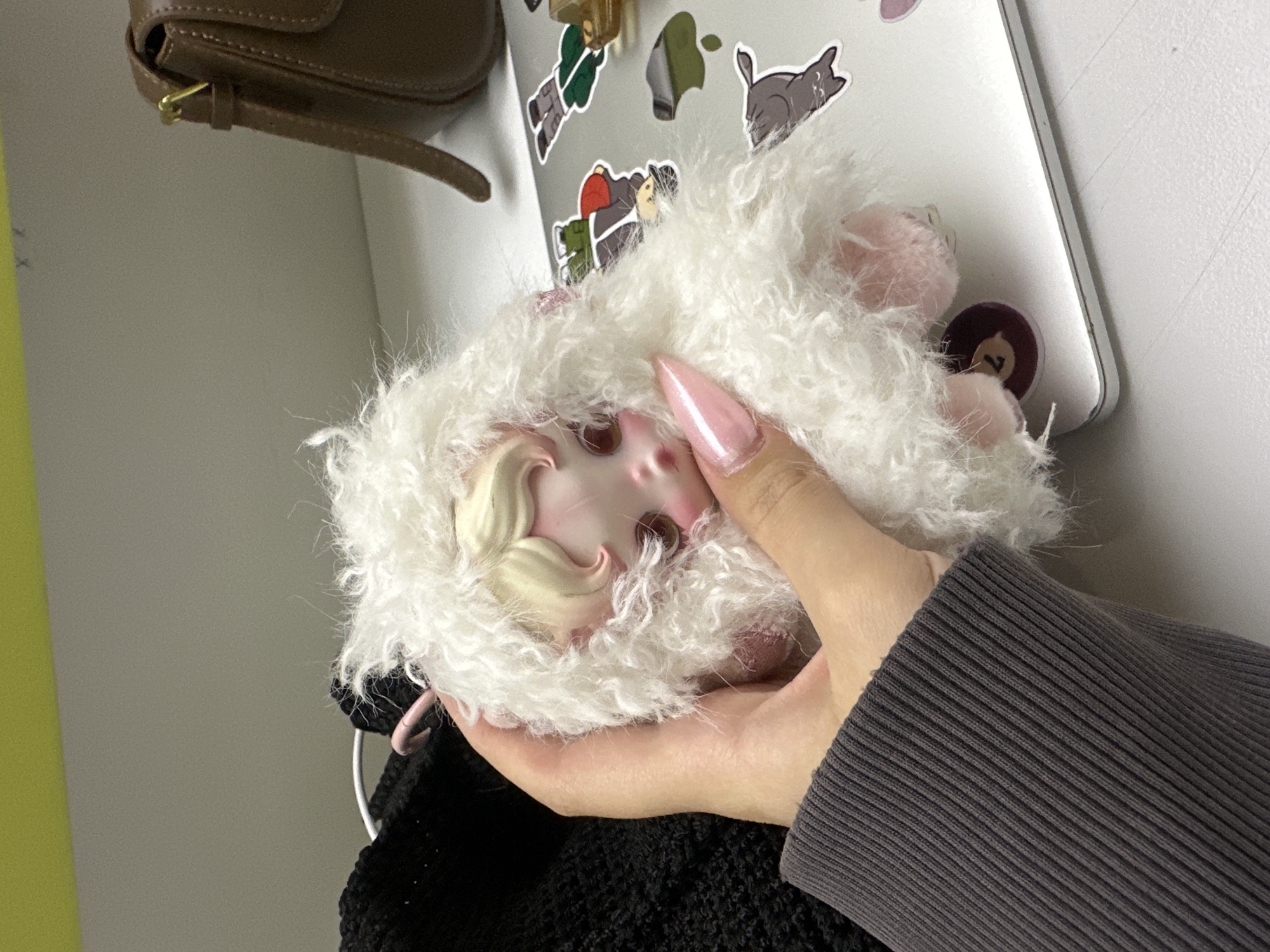 The image presents a close-up view of a doll head with fluffy white hair, held in a hand. The doll has pale skin, light pink cheeks, and blonde hair styled in a curl near the forehead. The hand holding the doll has a long, pointed, light pink nail. The sleeve of a grey knit sweater is visible on the arm.
Behind the doll and hand, there is a light-colored, possibly silver, surface covered in stickers. Some of the stickers are small, colorful figures.
The background suggests an indoor setting with a white wall on the right and a portion of a light yellow wall to the left. A brown bag is partially visible in the upper left corner.
No information suggests this image was taken in Yerevan, Armenia.