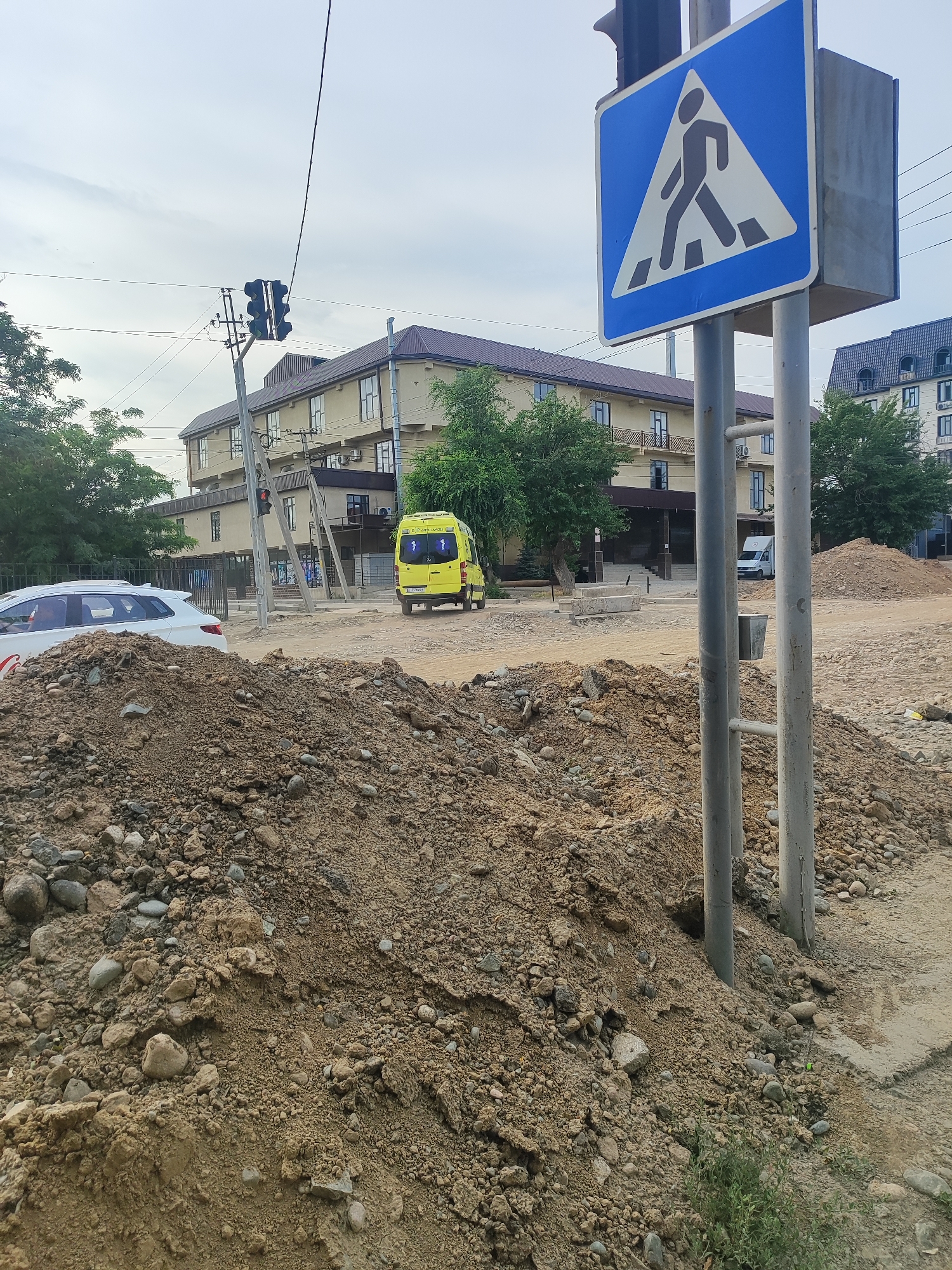 A daylight scene in Bishkek, Kyrgyzstan, shows a traffic intersection under construction. A blue and white pedestrian crossing sign is prominently displayed on a pole. Behind the sign, a traffic light is visible, with a yellow ambulance parked in the background. The foreground is dominated by piles of dirt and gravel, indicative of ongoing construction work. Buildings with multiple stories line the street, with trees and foliage interspersed. The sky is overcast.