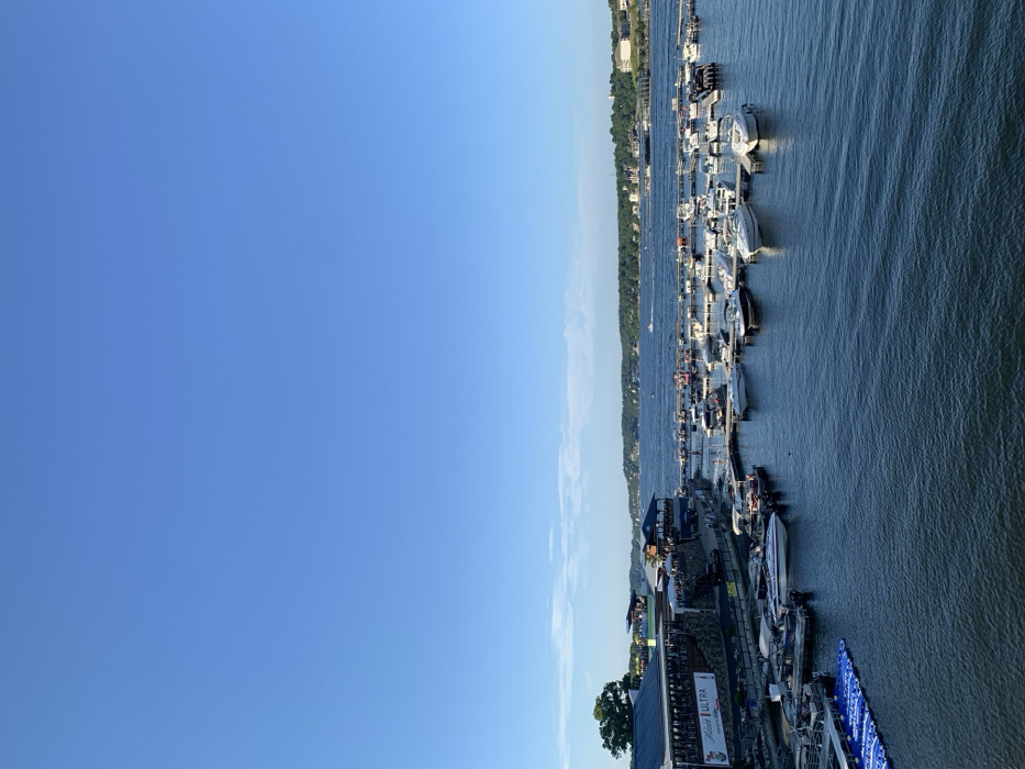 The image displays a clear blue sky over a large body of water, specifically a lake, located in Osage Beach, United States. The upper two-thirds of the frame consist of a bright, cloudless sky, with a faint horizontal band of wispy clouds near the horizon on the right. The lower portion features dark blue water with minor surface undulations.
On the left, a multi-story commercial building with a blue roof and a stone-clad base is visible, featuring a large "Michelob ULTRA" sign on its side. Several small, indistinct human figures are present on the outdoor patios of this establishment and on the adjacent piers. Extending from the shoreline into the water is a marina system, occupied by numerous recreational boats, predominantly white-hulled cruisers and pontoon boats, moored to light-colored floating docks. A bright blue modular floating dock is partially visible in the immediate lower-left foreground.
The far shoreline across the lake is densely forested with green trees, interspersed with various residential or commercial structures. Multiple small boats, possibly powerboats or personal watercraft, are visible in motion on the distant water. The scene indicates fair weather conditions during daylight hours in a recreational lake environment. No direct interactions or specific actions of the distant human figures are discernible.