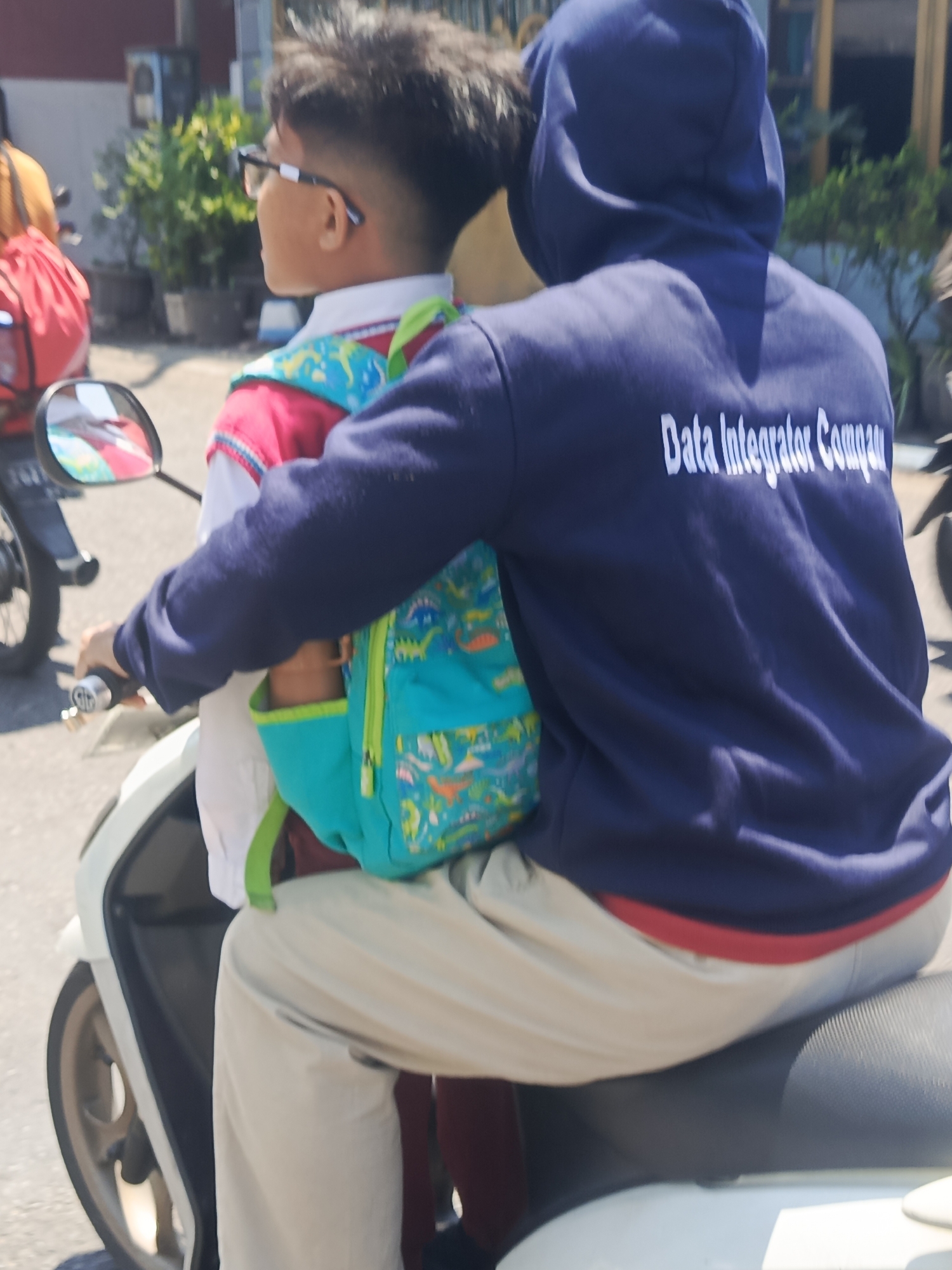 Two individuals are riding a white scooter on a paved road. The person in the front, a young boy, is wearing a white shirt with a red collar and a blue backpack with a dinosaur pattern. He has short, dark hair and is wearing glasses. The person in the back, an adult, is wearing a dark blue hooded sweatshirt with white lettering that reads "Data Integrator Company" and light-colored pants. The adult's arm is around the boy. In the background, there are buildings and some greenery. The lighting suggests it is daytime. The scene appears to be in Taman, Indonesia, based on the provided information.