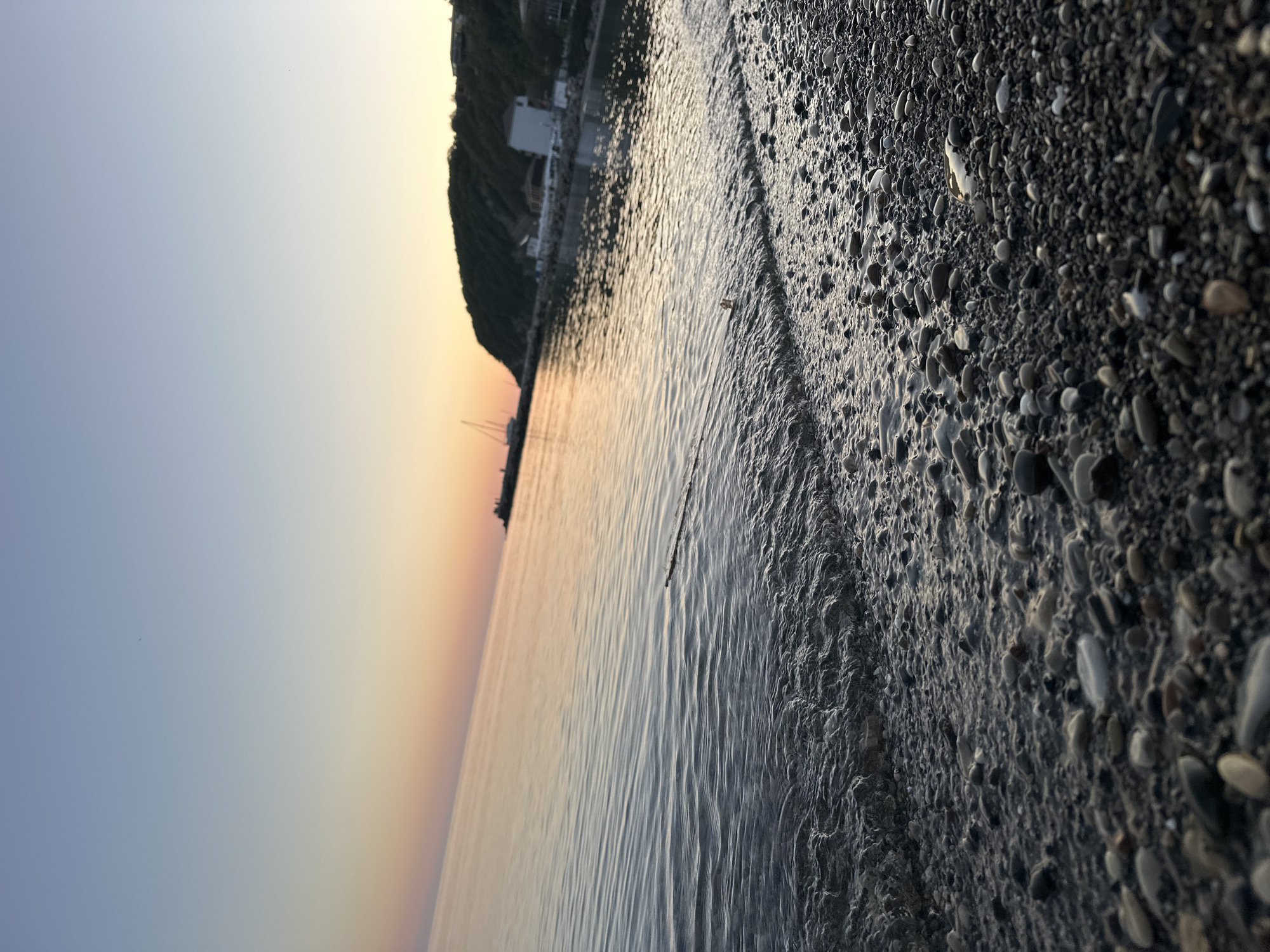 The image depicts a pebble beach in Novomikhaylovsky, Russia, at sunset. The sky transitions from blue to orange near the horizon, which is reflected in the calm water. The shore is covered in pebbles of varying sizes and colors, some wet from the receding waves. In the distance, a dark, vegetated cliff rises, with a white building visible on its slope. A sailboat is silhouetted against the sunset.