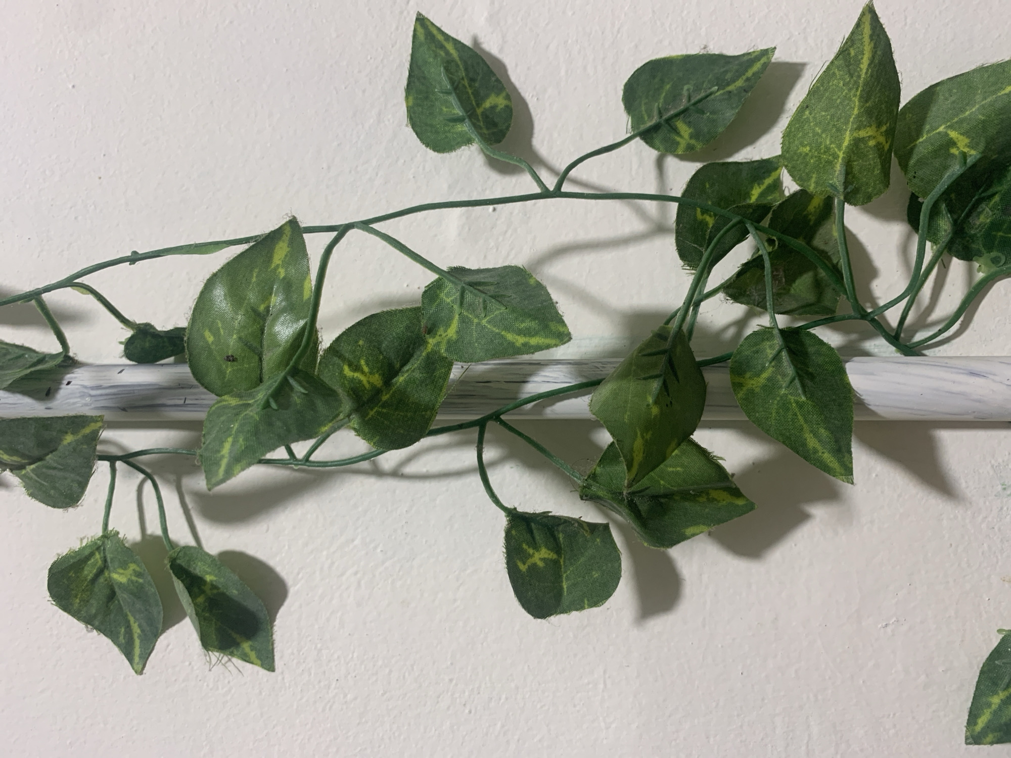 A close-up, eye-level shot shows a white pole with a marbled pattern partially visible on the left, serving as a support for a green artificial vine. The vine has numerous heart-shaped leaves with yellow variegation. The leaves are attached to thin green stems that twist and turn around the pole and extend towards the right side of the frame. The background is a textured, off-white wall, casting soft shadows from the vine. The lighting appears to be from the left, creating subtle highlights and shadows on the leaves and the wall. There are no people or animals visible in the image. The scene is indoors or under a covered area, given the controlled lighting and textured wall which is typical of interior walls in homes or buildings. The specific location in Kuala Pembuang, Indonesia, cannot be determined from the visual information alone, as the image depicts a common decorative element found in many settings globally.