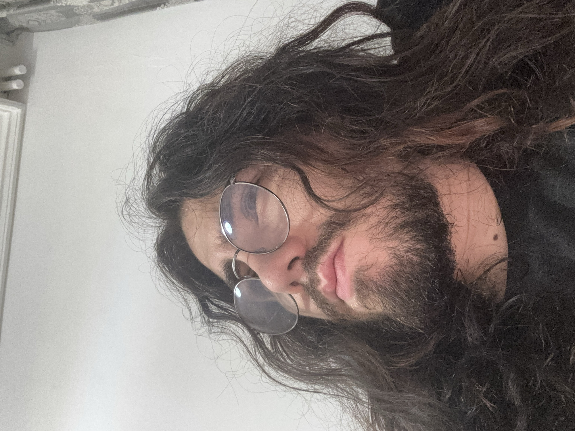 An extreme close-up, low-angle shot shows a person's face, from the nose down, with long, dark, messy hair framing the head. The person is wearing round, wire-rimmed glasses and has a beard and mustache. The lips are slightly parted. The background is a plain, light-colored wall with decorative molding at the top. The lighting is bright and evenly distributed.