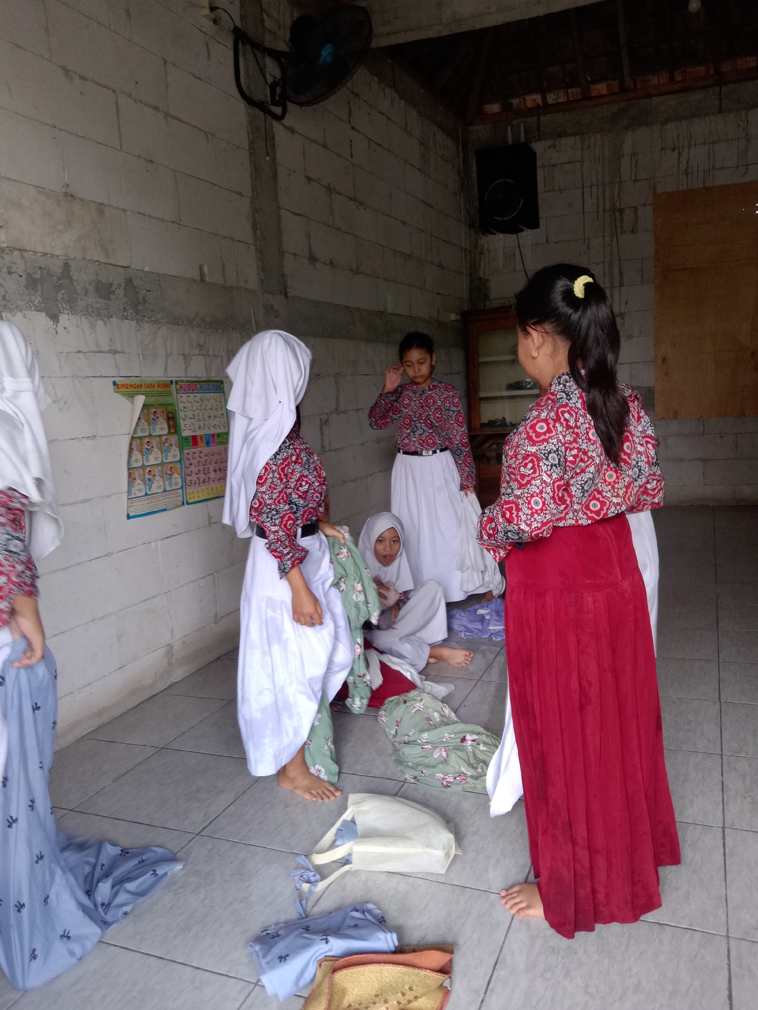 In Sugihwaras, Indonesia, a group of young girls in modest Islamic attire are gathered in an indoor, open-air space with rough brick walls and a wooden ceiling. The scene appears to be a communal or educational setting, possibly a madrasah or a community hall. Several girls are dressed in white hijabs and long, flowing white skirts. Some wear patterned shirts with red and white floral designs, while others have simpler white tops. One girl in the foreground wears a vibrant red skirt and a matching red and white patterned top, her hair pulled back in a ponytail. Another girl, sitting on the floor, wears a light green patterned skirt and a white top, her hijab framing her face. They seem to be in the process of changing or preparing for an activity. Clothing items, including patterned skirts and a light blue fabric with a dark pattern, are scattered on the tiled floor, alongside a white tote bag and what looks like a woven mat. A poster with colorful images and text, possibly educational material in Indonesian, is displayed on the brick wall. A black electric fan is mounted on the wall, and a speaker is visible above it. The lighting suggests it is daytime, with light filtering into the space. The overall impression is one of a casual gathering, perhaps during a break or transition in their daily routines.