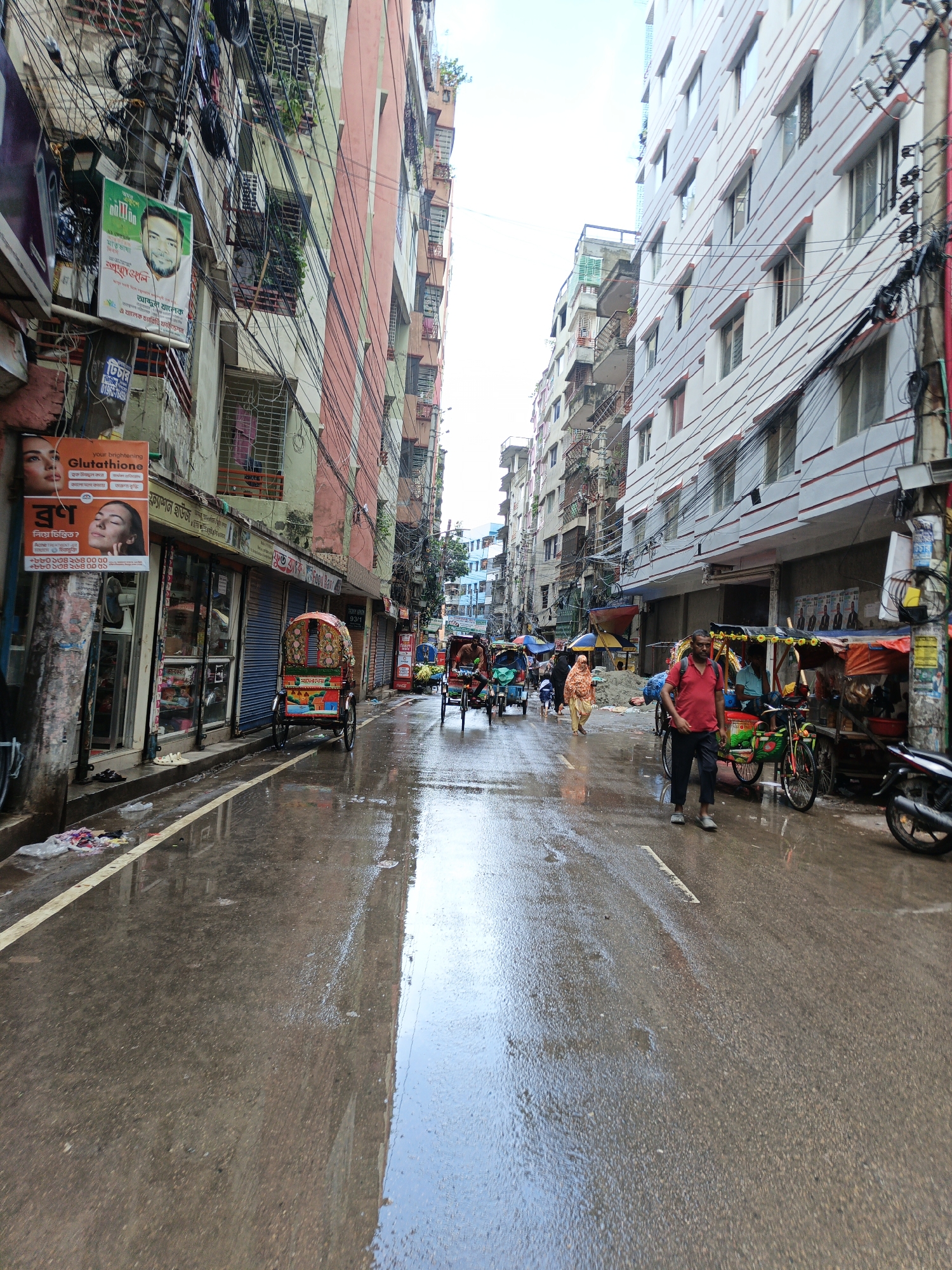 A street in Dhaka, Bangladesh, is wet and reflects the surrounding buildings, suggesting it has recently rained. Tall, multi-story buildings line both sides of the narrow street, with the left side featuring older, colorful buildings covered in a dense network of electrical wires. The right side has a more modern building with white siding and red trim. Several cycle rickshaws, some with colorful canopies, are present on the street, with some parked and others in motion. A few people are visible, including a man in a red shirt walking down the street, a woman in a headscarf, and individuals in rickshaws. Shops with closed shutters are also visible along the street. Advertisements, including one for "Glutathione," are posted on the buildings. The scene depicts a typical busy urban street in Dhaka.