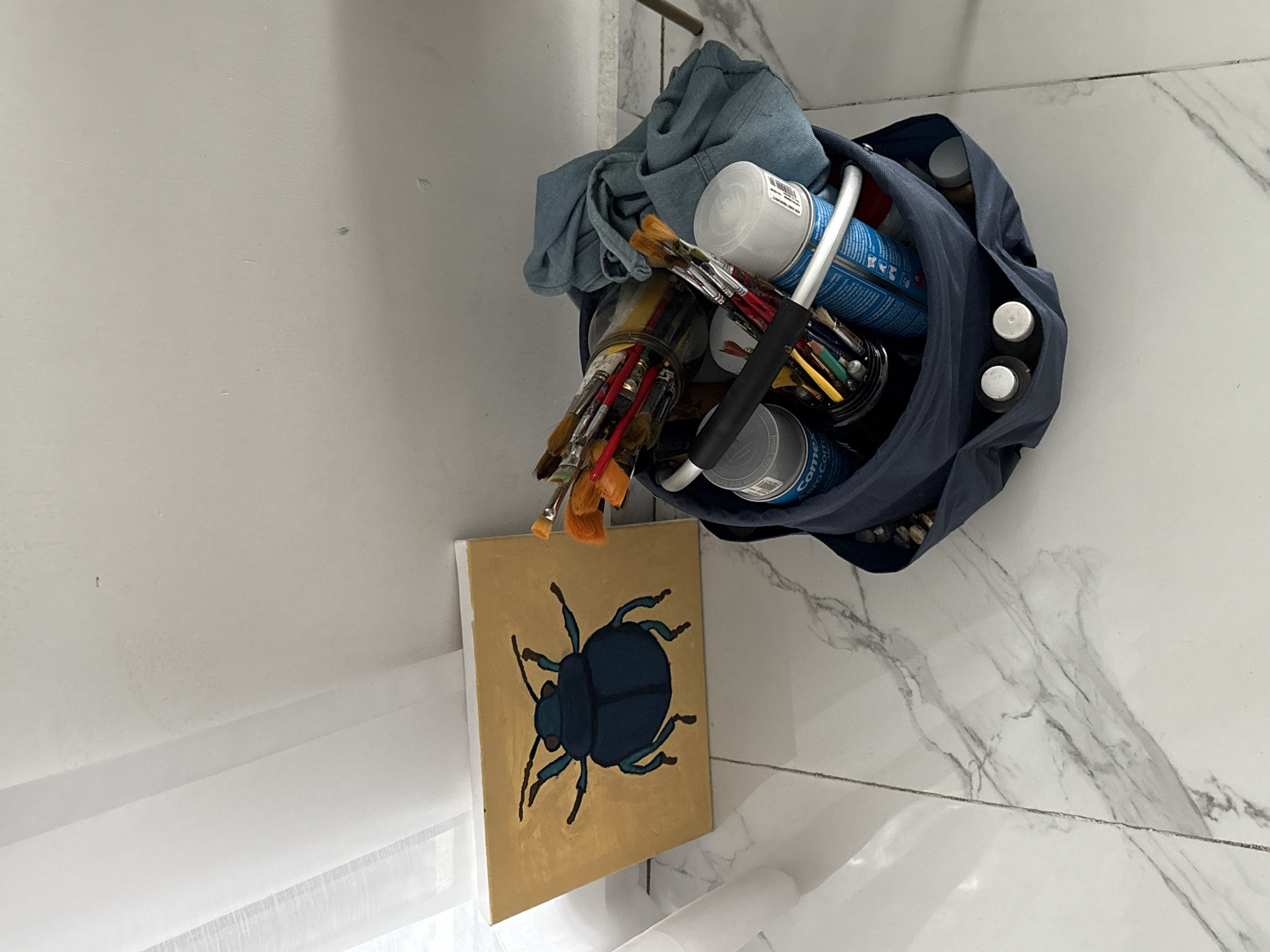 A painting of a blue beetle on a gold background leans against a white wall. To the right of the painting is a dark blue fabric bag filled with art supplies, including paintbrushes, spray cans, and small bottles of paint. The items are on a marble-tiled floor, with sheer white curtains visible on the left. The scene is located indoors, possibly in a studio or art space. The specific location of San Bernardino Tlaxcalancingo, Mexico, cannot be determined from the visual information provided in the image.