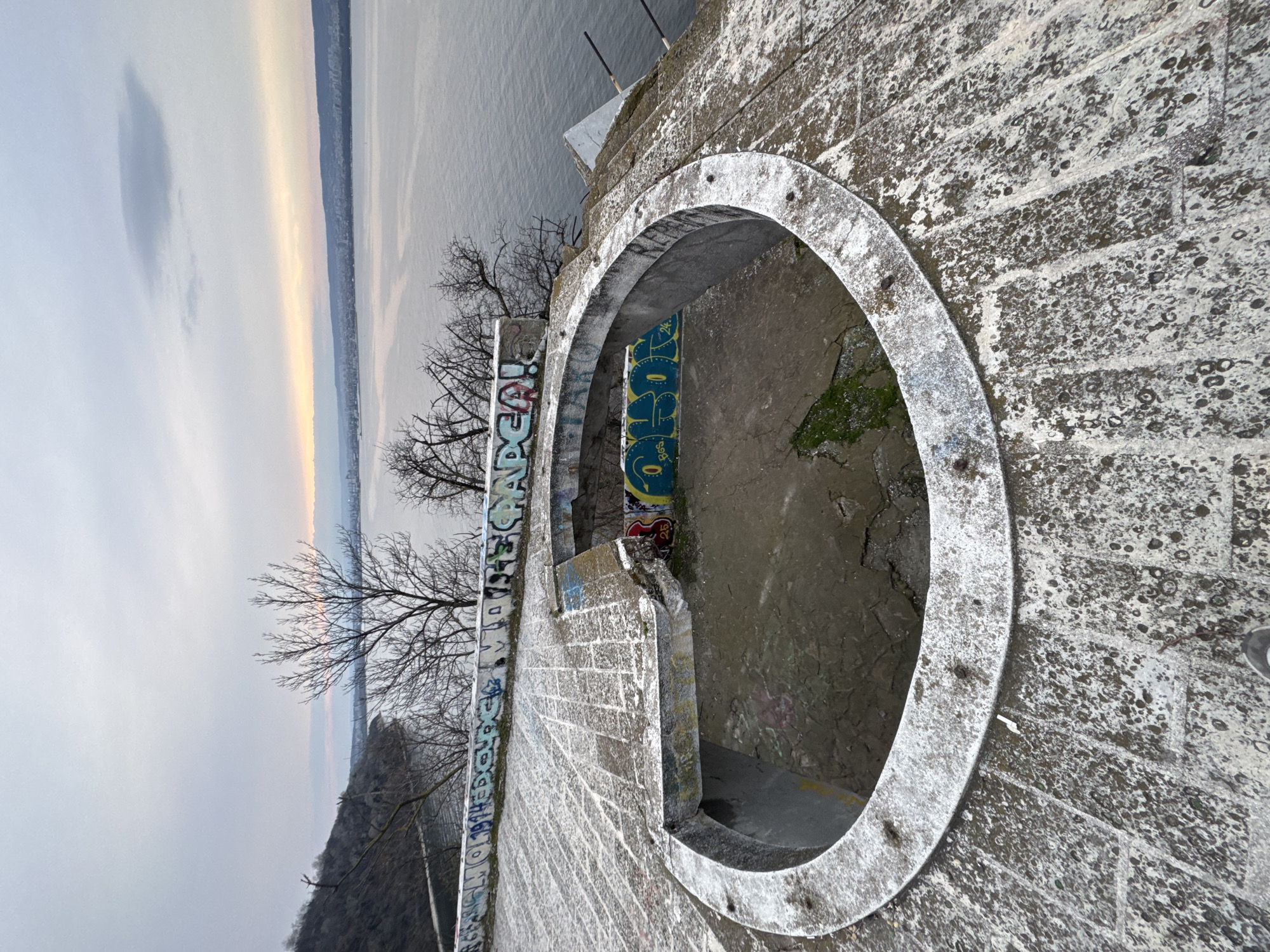 A high-angle shot captures a circular concrete structure, possibly a former observation point or part of a larger architectural complex. Graffiti adorns the interior and exterior walls of the structure. Bare trees are visible nearby. Beyond the structure, a body of water, likely the Black Sea, stretches into the horizon under a cloudy sky. The image appears to be taken in Varna, Bulgaria, based on the architectural style and geographic location. The environment seems decaying and abandoned, suggested by the graffiti and the deteriorating condition of the concrete. No people are visible in the frame.