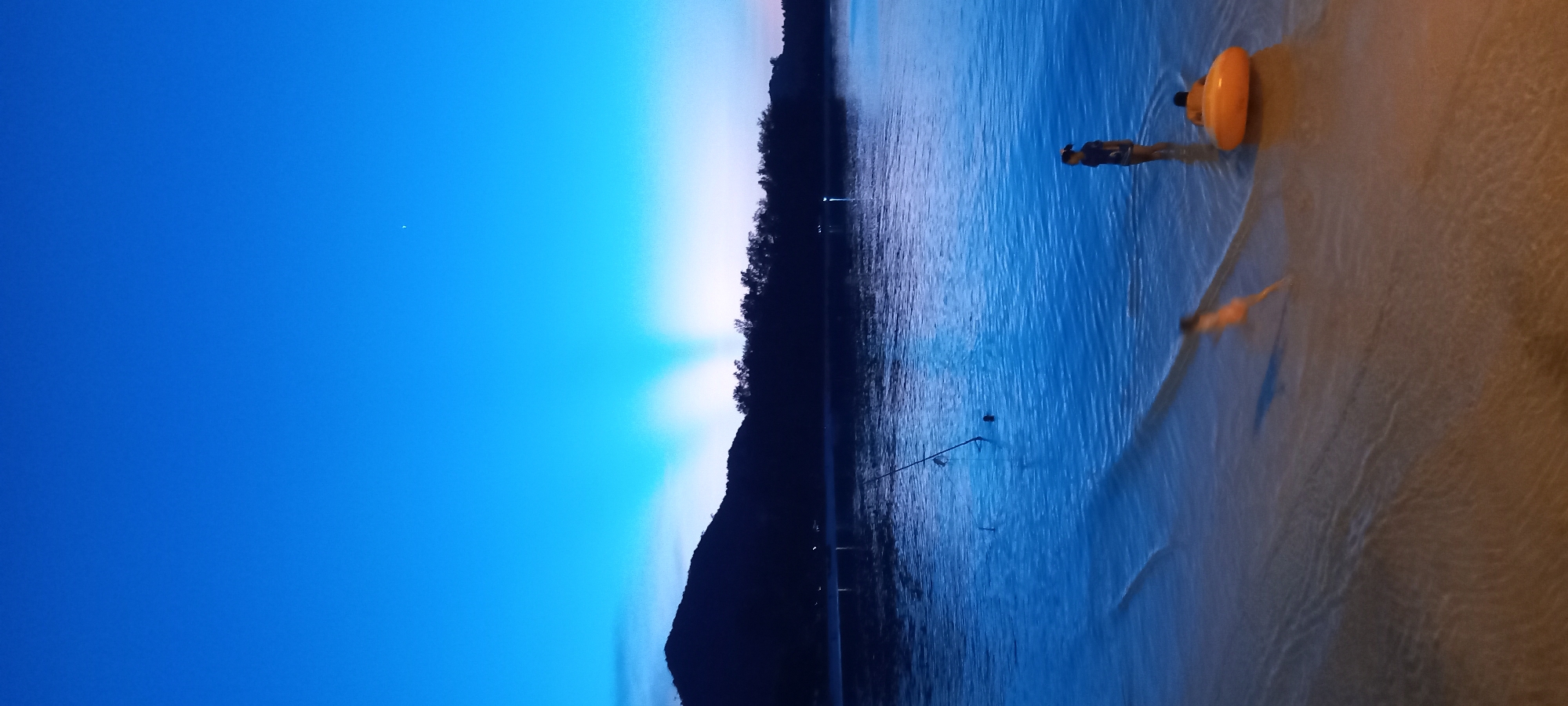 This image captures a serene coastal scene at either dawn or dusk in Baan Chaloklum, Thailand. The sky, dominating the upper portion, transitions from a deep, vibrant blue at the top to a lighter blue and soft white, tinged with pink, near the horizon. Prominent crepuscular rays fan out from behind a distant, dark landmass, which is densely covered with trees and appears mountainous. Small lights are visible along this distant shore, hinting at a settlement or infrastructure.
In the foreground, calm, shallow water meets a gently sloping, wet sandy beach. A person, likely a child, stands in the ankle-deep water, holding an orange inflatable ring. Their reflection is faintly visible on the wet sand. Further into the water, a blurred, smaller figure is discernible, possibly another person. A fishing rod is positioned in the water, extending diagonally from the middle ground, indicating fishing activity. The water's surface shows gentle ripples, reflecting the ambient light. A tiny white speck, possibly the moon or a bright star, is visible in the deep blue expanse of the sky. The overall atmosphere is peaceful and tranquil, bathed in the soft, blue-dominant light of early morning or late evening. No text is visible in the image.