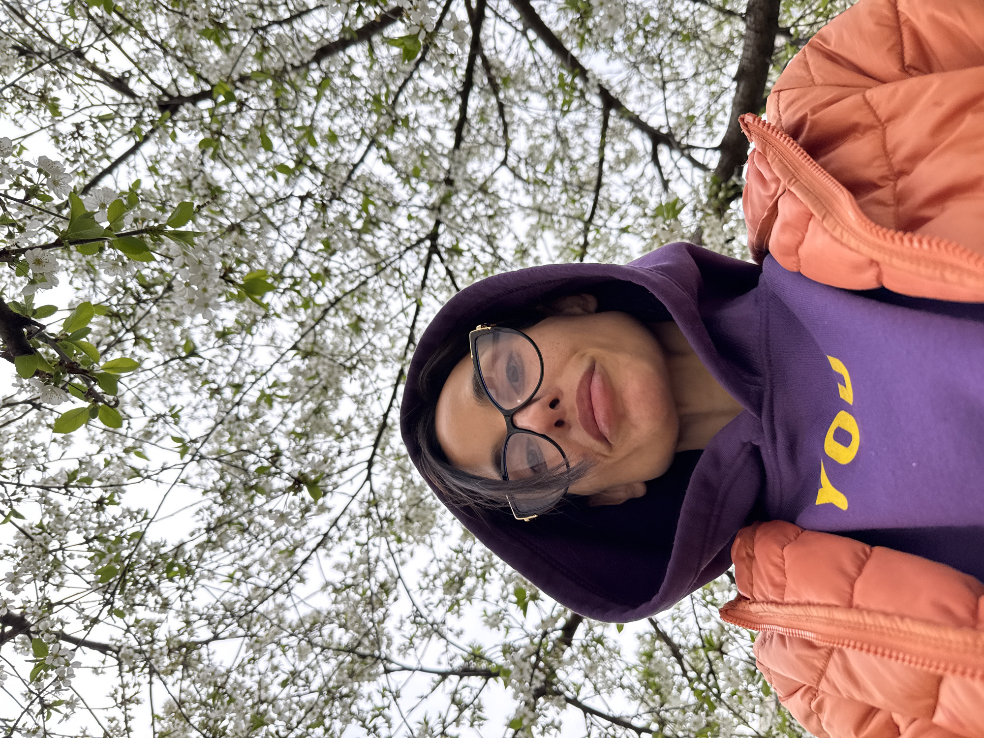 A woman wearing a purple hooded sweatshirt and an orange puffer jacket is standing beneath a tree in full bloom with white flowers. The woman is wearing glasses. The photograph is taken from a low angle, looking up at the woman and the tree. The sweatshirt has a yellow "You" logo. The location is identified as Pleven, Bulgaria.