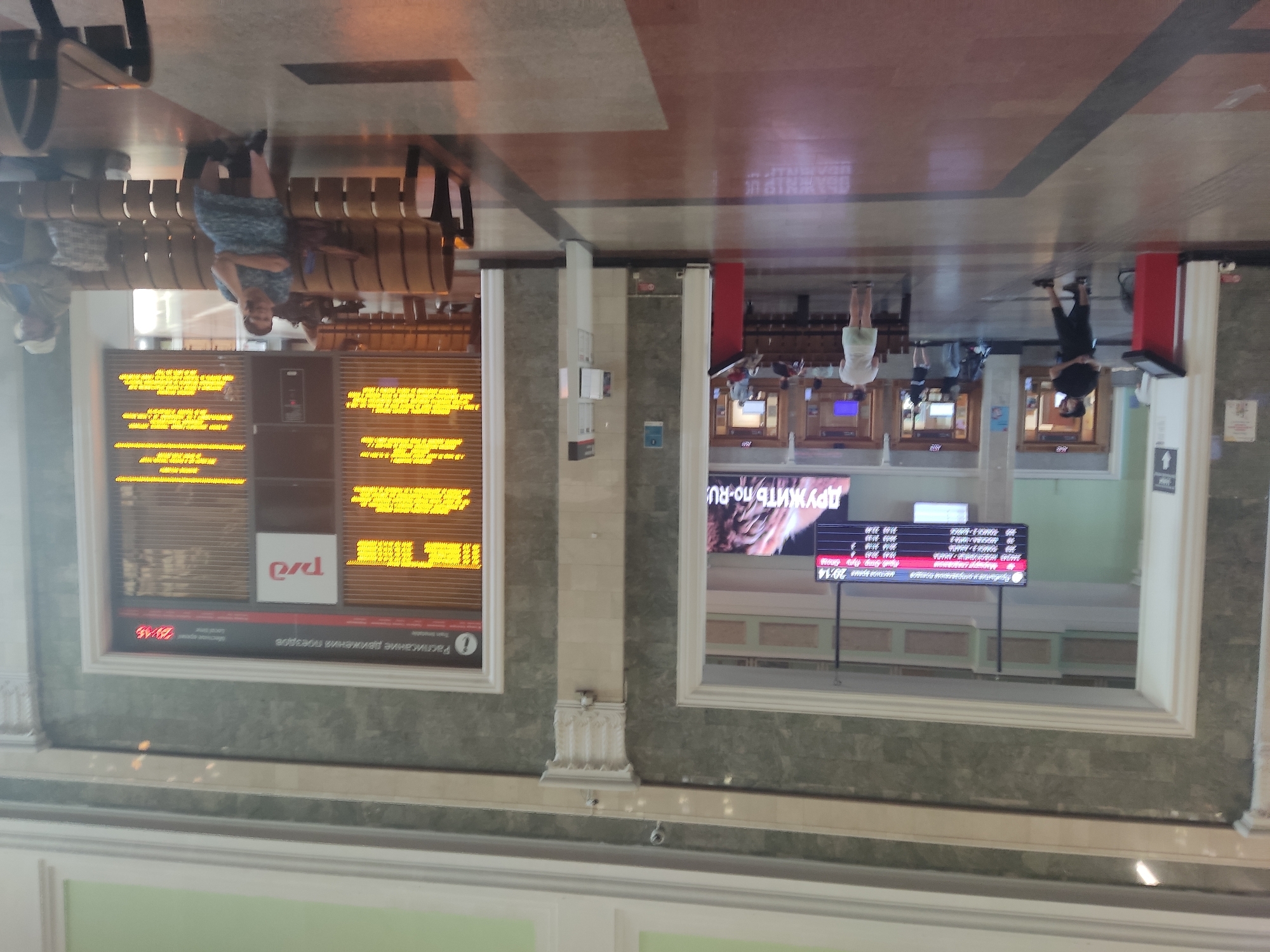 The image shows the interior of a train station. In the foreground, there are patterned floor tiles. In the midground, there are several people standing and sitting on benches. To the left, a digital display board shows train departure and arrival information. To the right, another large digital display board shows train schedules in yellow text. The architecture includes marble-patterned walls and decorative columns. This appears to be a waiting area for passengers. The presence of the "РЖД" logo, which stands for Russian Railways, confirms it's a Russian railway station. Given the style and the context of the Russian Railways logo, this is likely the Novosibirsk-Glavny railway station in Novosibirsk, Russia.