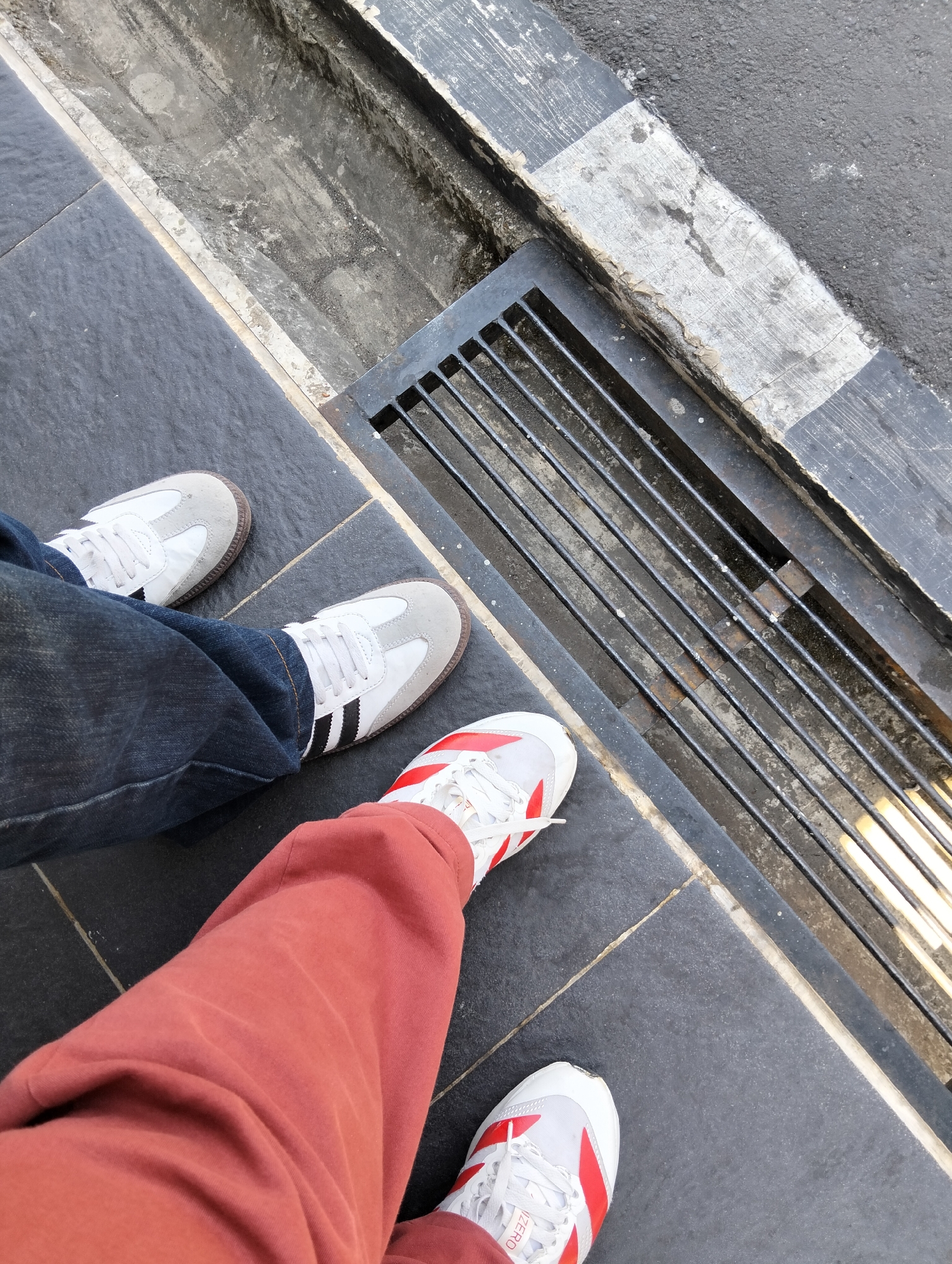 Two pairs of feet are visible, standing on a dark tiled surface next to a drainage grate. The left pair of feet is wearing blue jeans and white and grey Adidas Samba sneakers. The right pair of feet is wearing rusty-orange colored pants and white sneakers with red accents. The drainage grate is made of black metal bars and is situated in a concrete trench. The background shows asphalt and the edge of a sidewalk with a weathered white and black painted line. The scene appears to be an outdoor urban environment, possibly near a street or walkway.