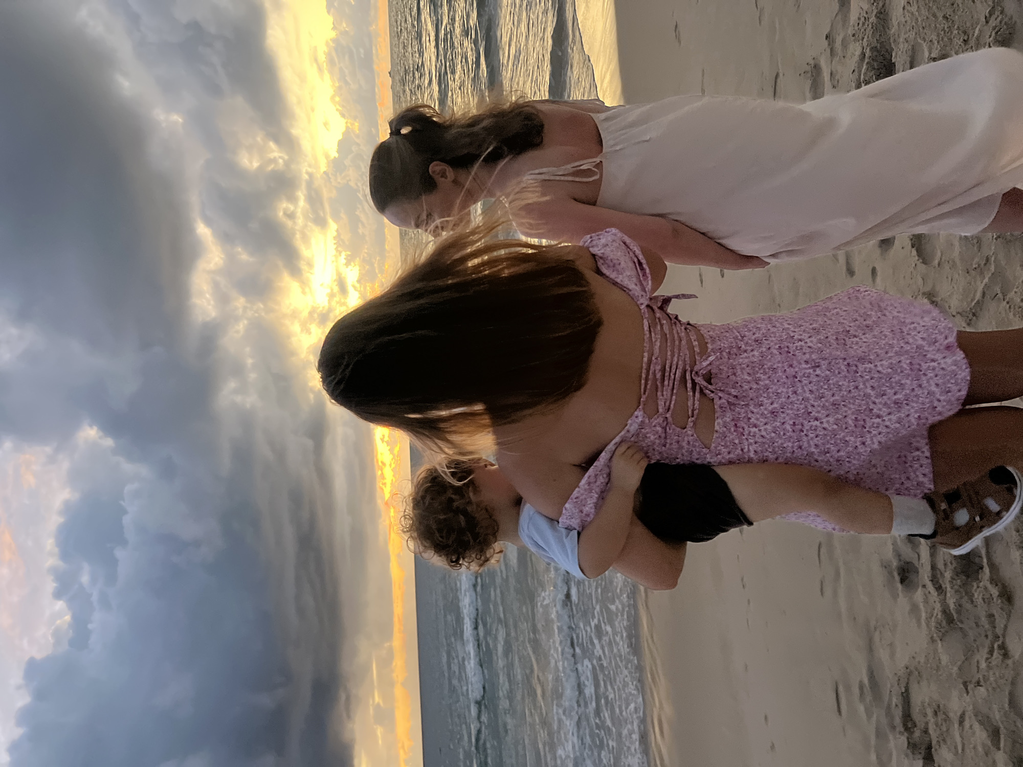 Three people stand on a sandy beach facing the ocean at sunset. A person in the foreground, viewed from the back, holds a young child in their arms. The child is wearing a light blue shirt and dark shorts, with tan sandals. The adult holding the child is wearing a pink floral dress with a lace-up back. To the right, another person stands facing the ocean, wearing a long, light-colored dress. The sky is filled with dramatic clouds, with the sun setting on the horizon, casting a warm glow on the scene. The ocean is calm with gentle waves. The image appears to have been taken on a beach in Choeng Thale, Thailand.