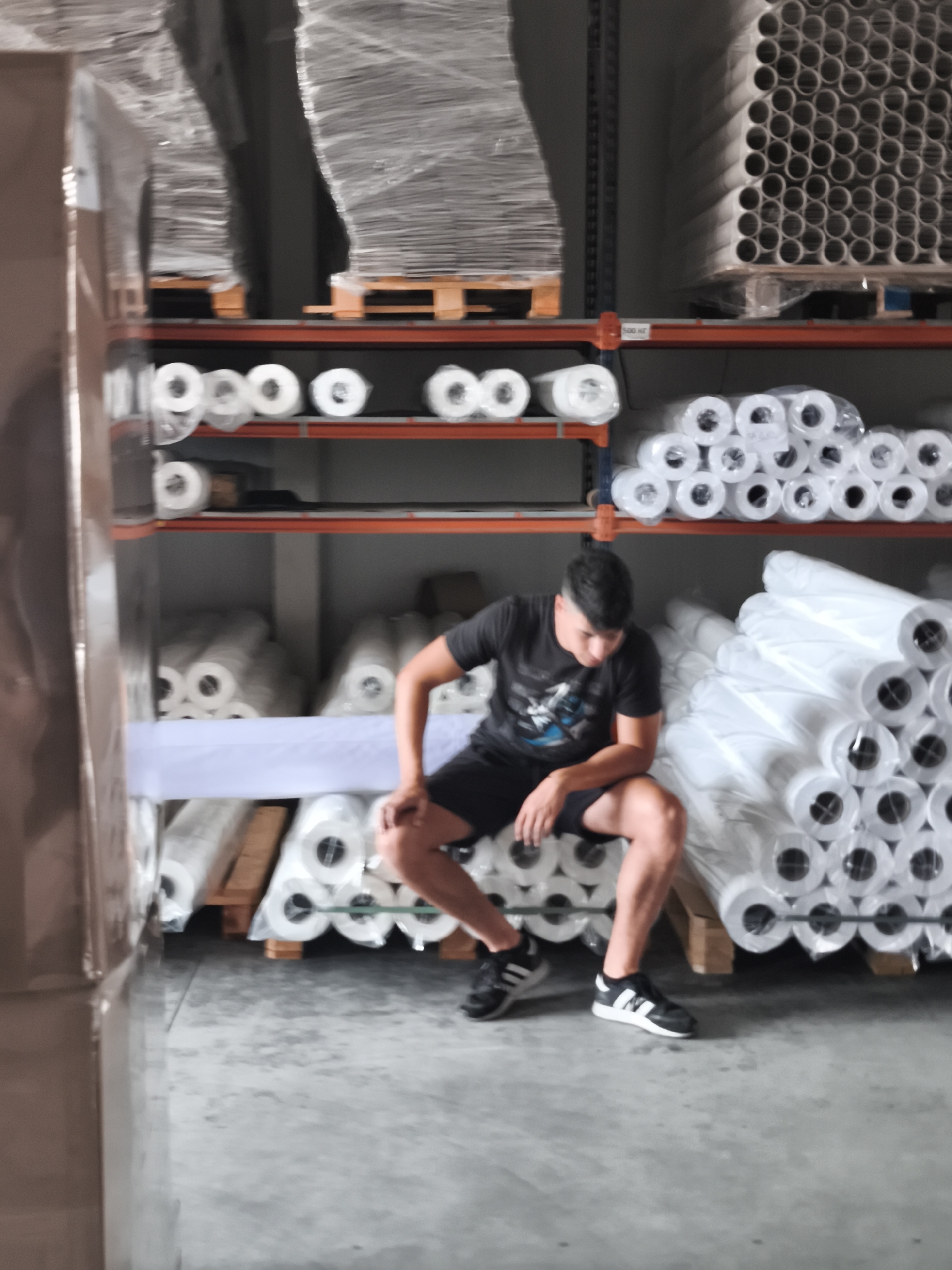 A young man with short dark hair is sitting on the floor in a warehouse, leaning back against a stack of large white rolls wrapped in plastic. He is wearing a black t-shirt with a graphic print and black athletic shorts, as well as black and white athletic shoes. His left leg is bent at the knee, and his right leg is extended slightly forward. His hands are resting on his lap. The warehouse shelves are filled with various rolls of material, some of which are also wrapped in plastic. In the background, large stacks of what appear to be paper or fabric rolls are visible on pallets. The floor is concrete.