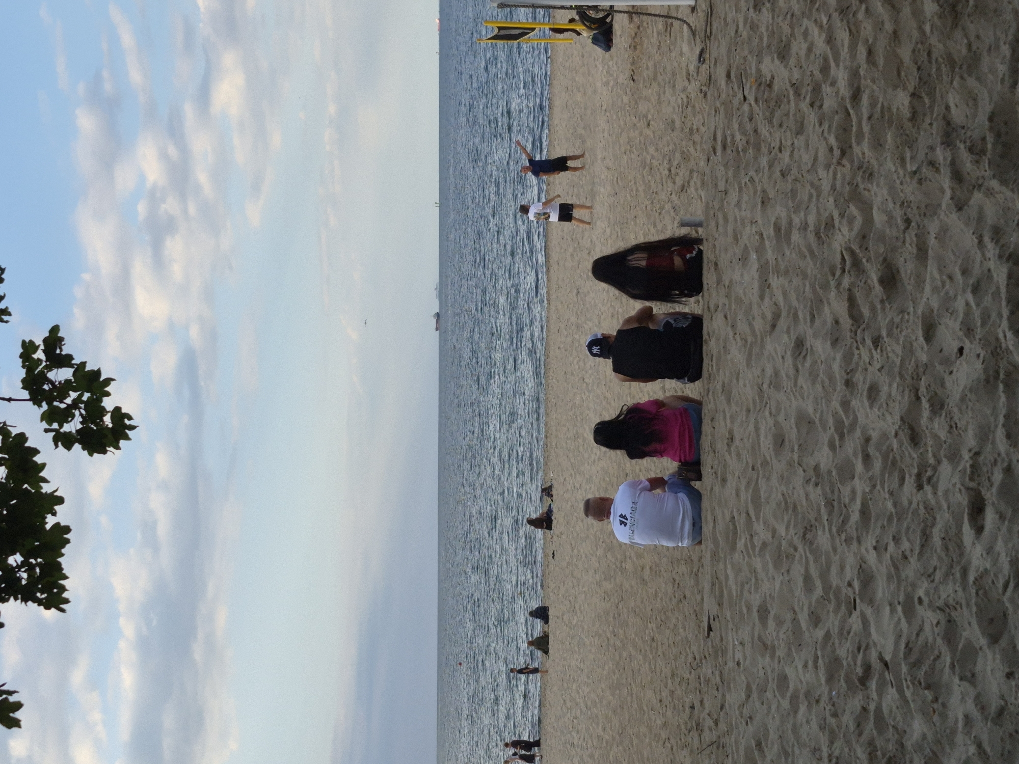 The image depicts a sandy beach with the ocean in the background. The scene is likely Gdańsk, Poland. Several people are present on the beach, engaged in various activities.
In the foreground, several individuals are sitting or kneeling on the sand. One person, a man, is wearing a white t-shirt with visible text that appears to read "BALENCIAGA" and is kneeling on the sand, facing away from the camera. Another person, a woman, is wearing a bright pink top and blue shorts, also kneeling. Behind them, another person with long dark hair is seated. Further down the beach, more people are visible in smaller groups, some appearing to be standing or sitting.
In the middle ground, two men are standing near the water's edge. One is wearing a dark t-shirt and shorts and is in a dynamic pose, perhaps stretching or dancing, with his arms outstretched. The other man is wearing a white t-shirt and dark shorts.
In the background, there is a yellow and black structure, possibly part of a beach volleyball net or a lifeguard stand. The ocean appears calm, with gentle waves. A few boats or ships are visible in the distance on the horizon, suggesting maritime activity. The sky is a pale blue with scattered white clouds, indicating fair weather. The lighting suggests it might be late afternoon or early evening, with soft light bathing the scene. The overall mood appears relaxed and casual, characteristic of a day at the beach.