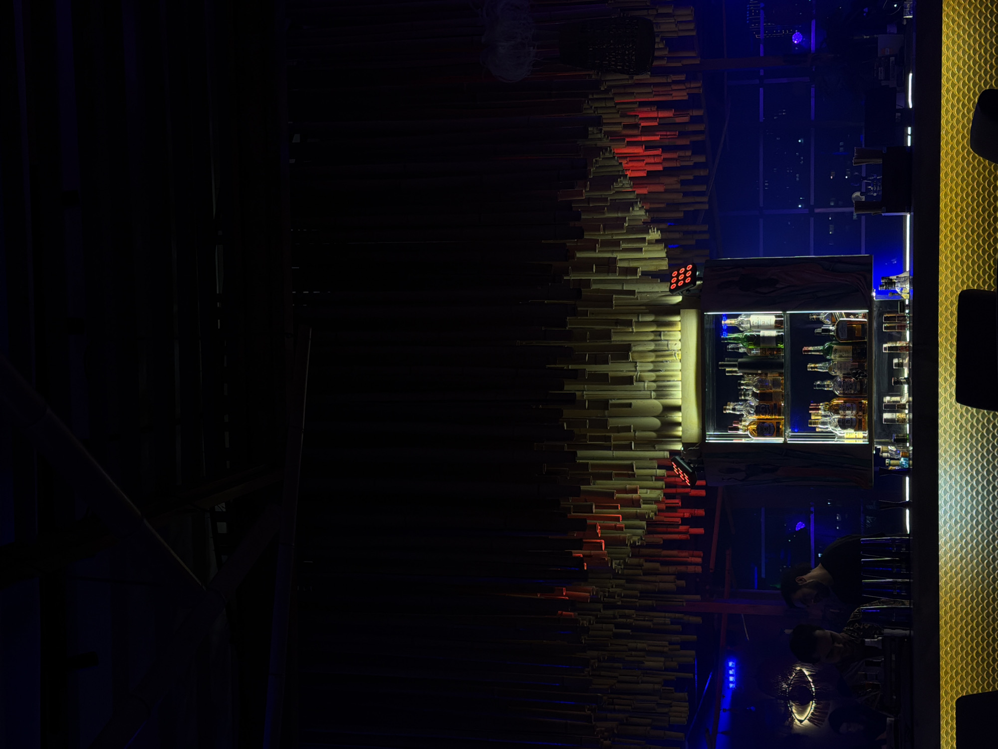 The image shows a bar area in a dimly lit establishment, likely in Yerevan, Armenia. The bar counter is on the right with a textured yellow surface and black seating. Behind the bar, a refrigerated display case is filled with bottles of alcohol. Above the display case, there's a decorative bamboo-like structure with red lighting. In the background, a window reveals a cityscape with blue lighting. Reflections of a person sitting at the bar are visible on the surface.