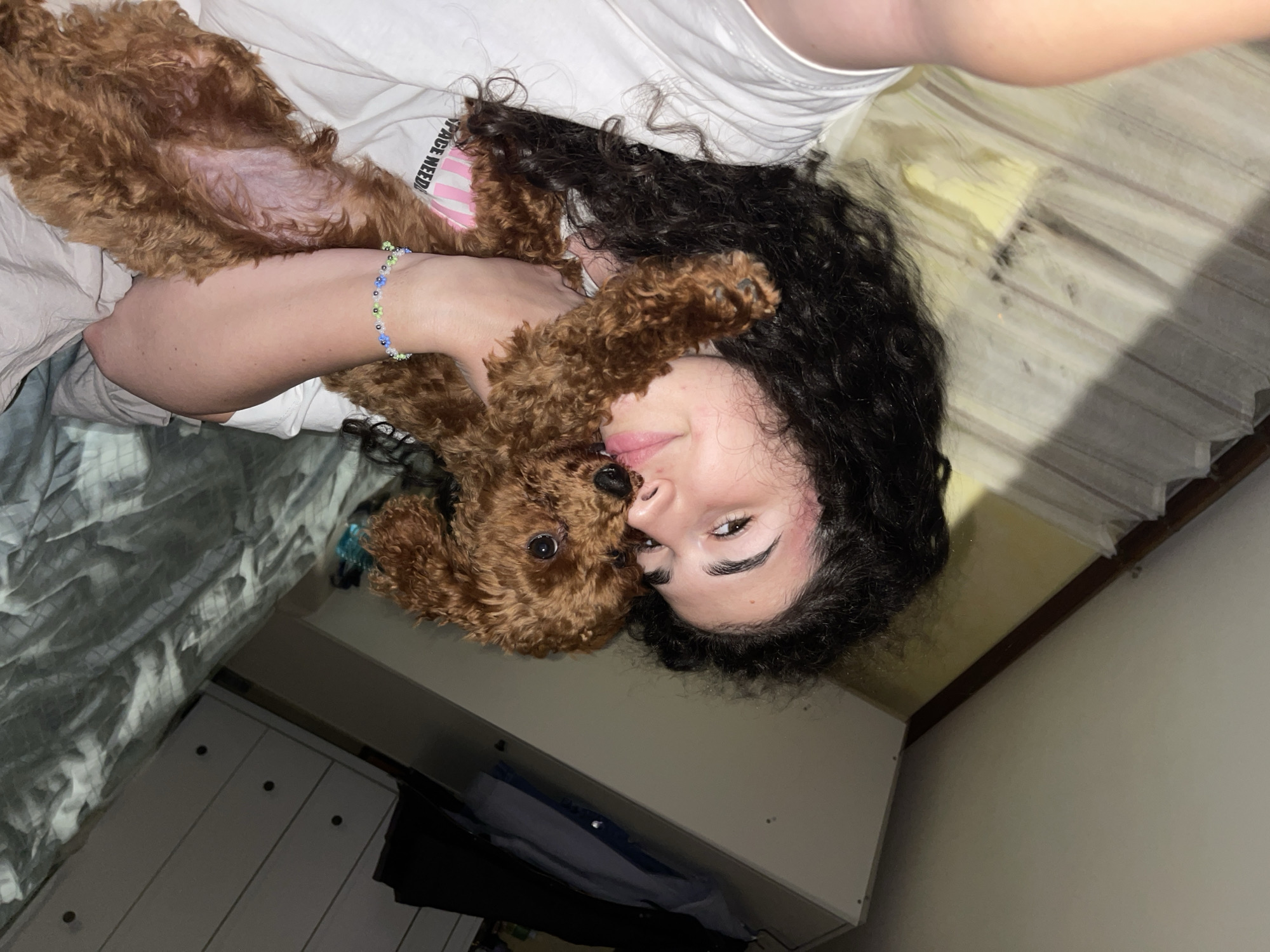The image depicts a person and a small dog cuddling intimately in an indoor setting, likely a bedroom in Sofia, Bulgaria. The photo is taken from an overhead perspective, with the subjects lying down.
The person, visible with dark, curly hair and a partial view of their face, appears to be smiling slightly. Their right arm, around which a colorful beaded bracelet with blue, green, and white floral designs is worn, is prominent. They are dressed in a white t-shirt.
A small, reddish-brown, curly-haired dog, resembling a poodle, rests on the person's chest and stomach. Its head is tucked close to the person's face. The dog is wearing a pink collar or harness, on which the text "SPACE WEED" is partially legible in white capital letters. A small shaved patch on the dog's belly is visible.
The scene is set in what appears to be a bedroom, indicated by the bedding: a white duvet cover with a subtle pattern beneath the person, and a yellow and light grey striped sheet near their head. In the bottom left, a white dresser or cabinet with dark knobs is partially visible, hinting at typical bedroom furniture. The lighting suggests an indoor environment, possibly softly lit or at night, contributing to a warm and affectionate ambiance between the person and their pet. The overall impression is one of comfort and close companionship.