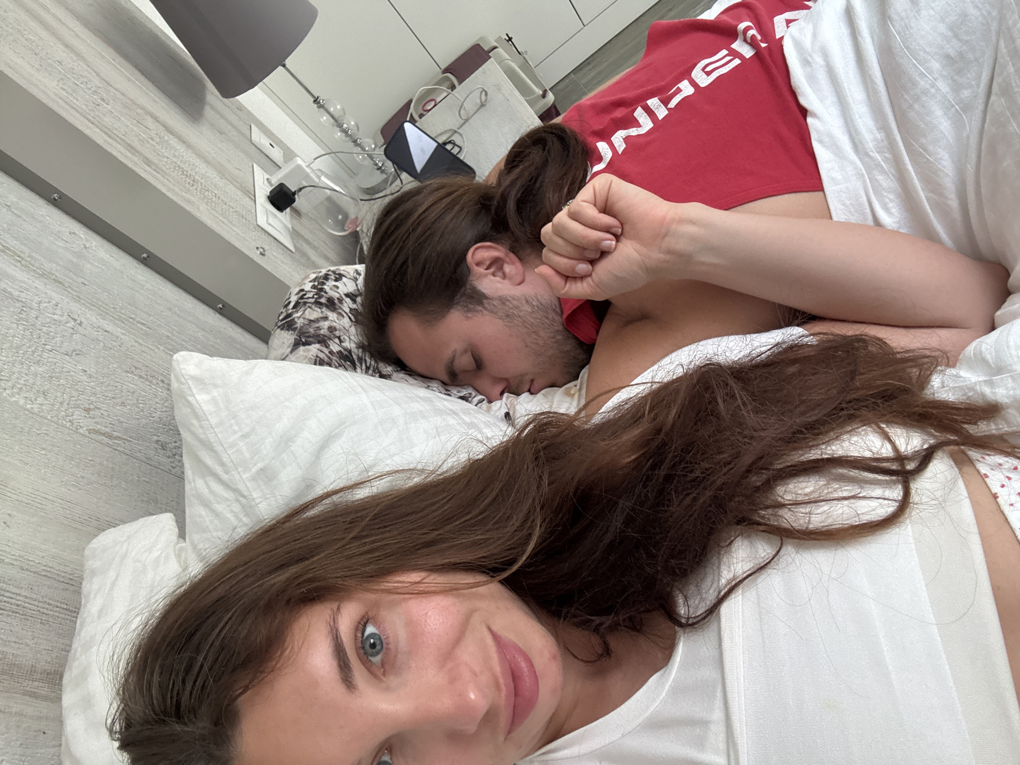 A close-up, slightly tilted selfie captures two individuals in a bedroom, depicting a peaceful morning or daytime scene in Valencia, Spain. In the foreground, a woman with long brown hair and striking blue eyes looks directly at the camera with a subtle, content smile. She is wearing a white sleeveless top.
Beside her, a man lies asleep, his face turned away from the camera. He has long brown hair tied in a ponytail and light facial stubble. He wears a red t-shirt with the word "UNDER" clearly visible across the back. His head rests on a patterned pillowcase, amidst white pillows and bedding.
The background reveals a modern bedroom interior. A light wood-grain headboard with metallic accents runs along the wall. On a bedside surface, a lamp with a gray shade and a decorative base is visible, alongside a white wall outlet with a black adapter plugged in. A small black and white electronic device, possibly a speaker, is also present. The overall ambiance is intimate and calm.