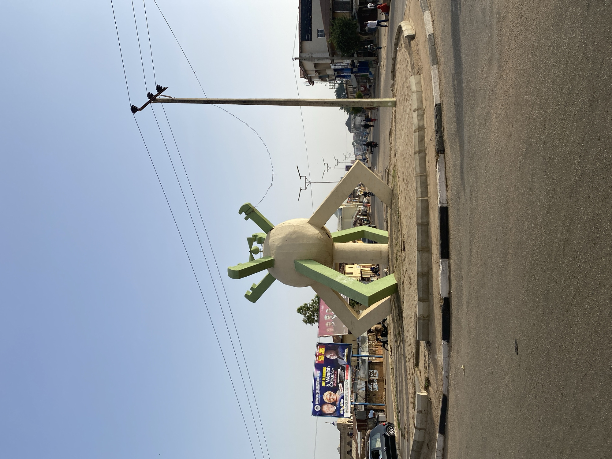 The image depicts a roundabout in Jos, Nigeria, featuring a spherical sculpture with green and beige appendages. The sculpture is situated on a concrete platform along the road's edge. Behind the sculpture, billboards with images of people and text advertising "Life Planning & Wealth Creation" are visible. A utility pole with power lines extends into the sky. The road is asphalt, and a sidewalk runs parallel to it. Buildings can be seen in the background. People and vehicles are present further down the road. The sky is clear and light blue.