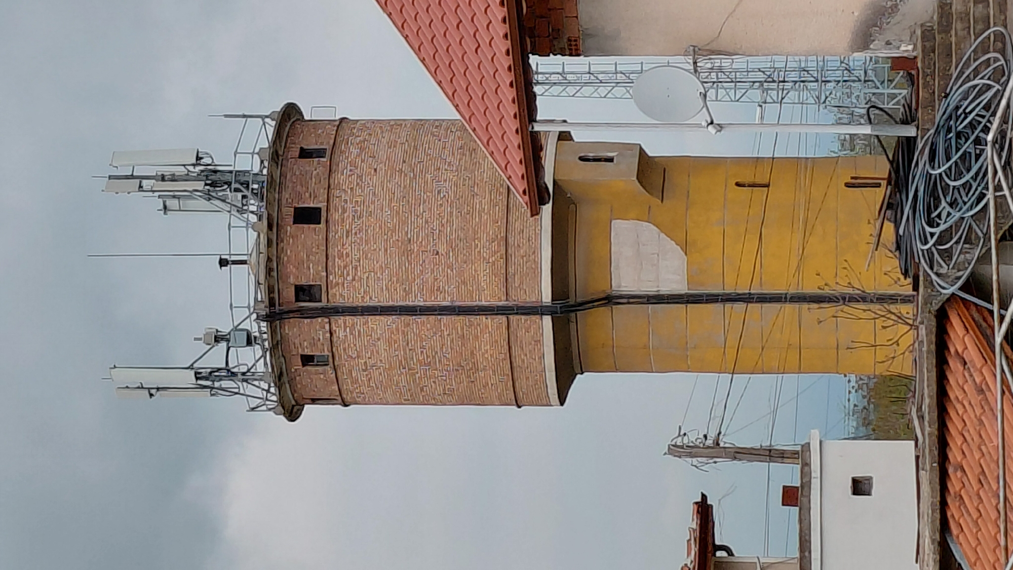 This image shows a two-story brick and yellow tower in Unknown, Unknown. The brick tower is topped with antennas. The yellow tower is adjacent to a building with a red tile roof. A satellite dish and coiled wires are visible on the roof of the building next to the yellow tower. Power lines are also visible in the background.