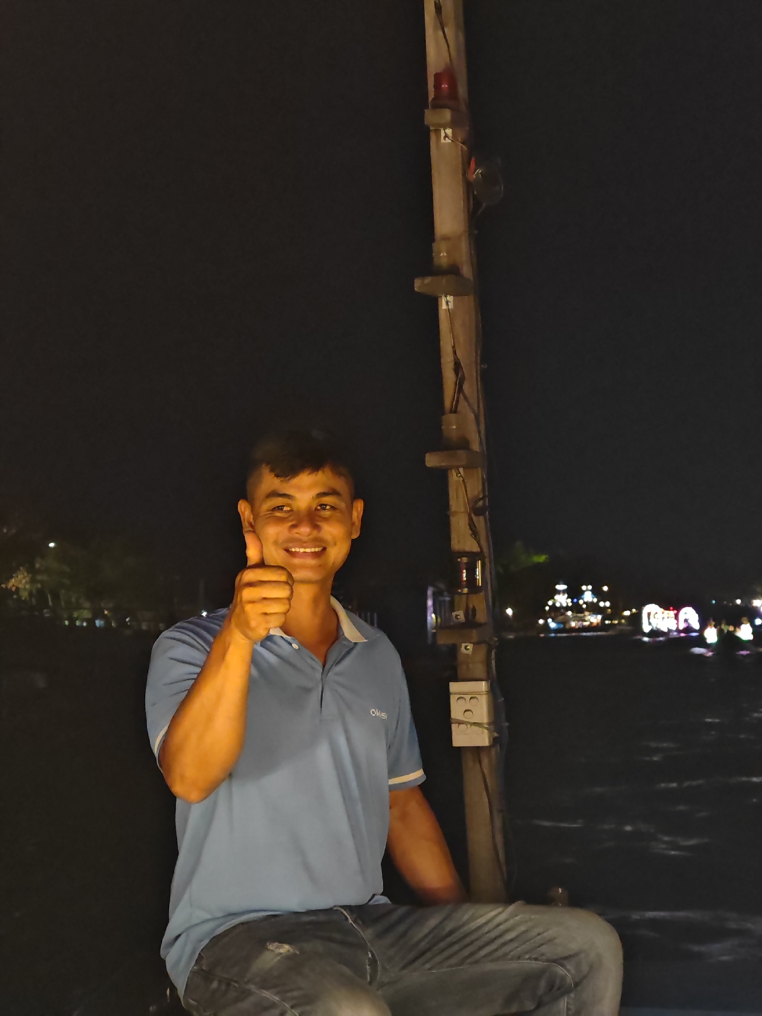 A man in a blue polo shirt and jeans gives a thumbs-up gesture. He is sitting on a boat at night, with the lights of Hoi An, Vietnam, visible in the background. The water reflects the colorful lights of the city. A wooden pole with electrical outlets and a red light is to the man's right.