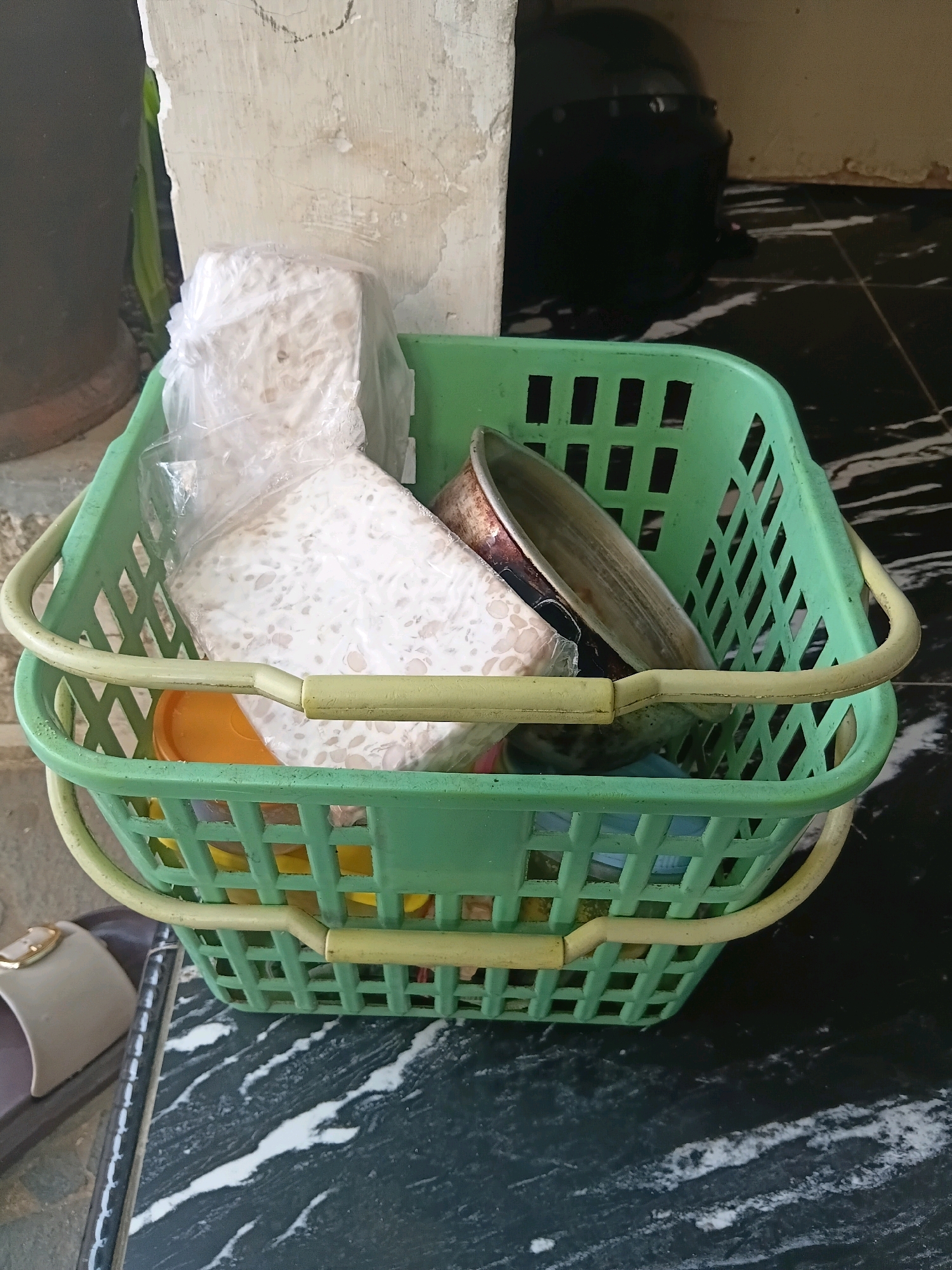 A pair of green plastic baskets, nested one inside the other, are positioned on a marble-patterned floor. The upper basket contains a large block of white, porous food product, likely tempeh, wrapped in clear plastic. Beneath the tempeh, an orange plastic container is visible, along with what appears to be a metal pan. Various other items are partially obscured within the lower basket. To the left of the baskets, a dark brown planter pot and a portion of a wall with distressed plaster are present. In the background, a dark object, possibly a motorcycle helmet, rests against the wall. A single sandal with a light-colored strap and a gold buckle is lying on the floor to the left of the baskets. The scene appears to be outdoors, possibly on a porch or walkway, given the presence of the wall and the floor tiling.