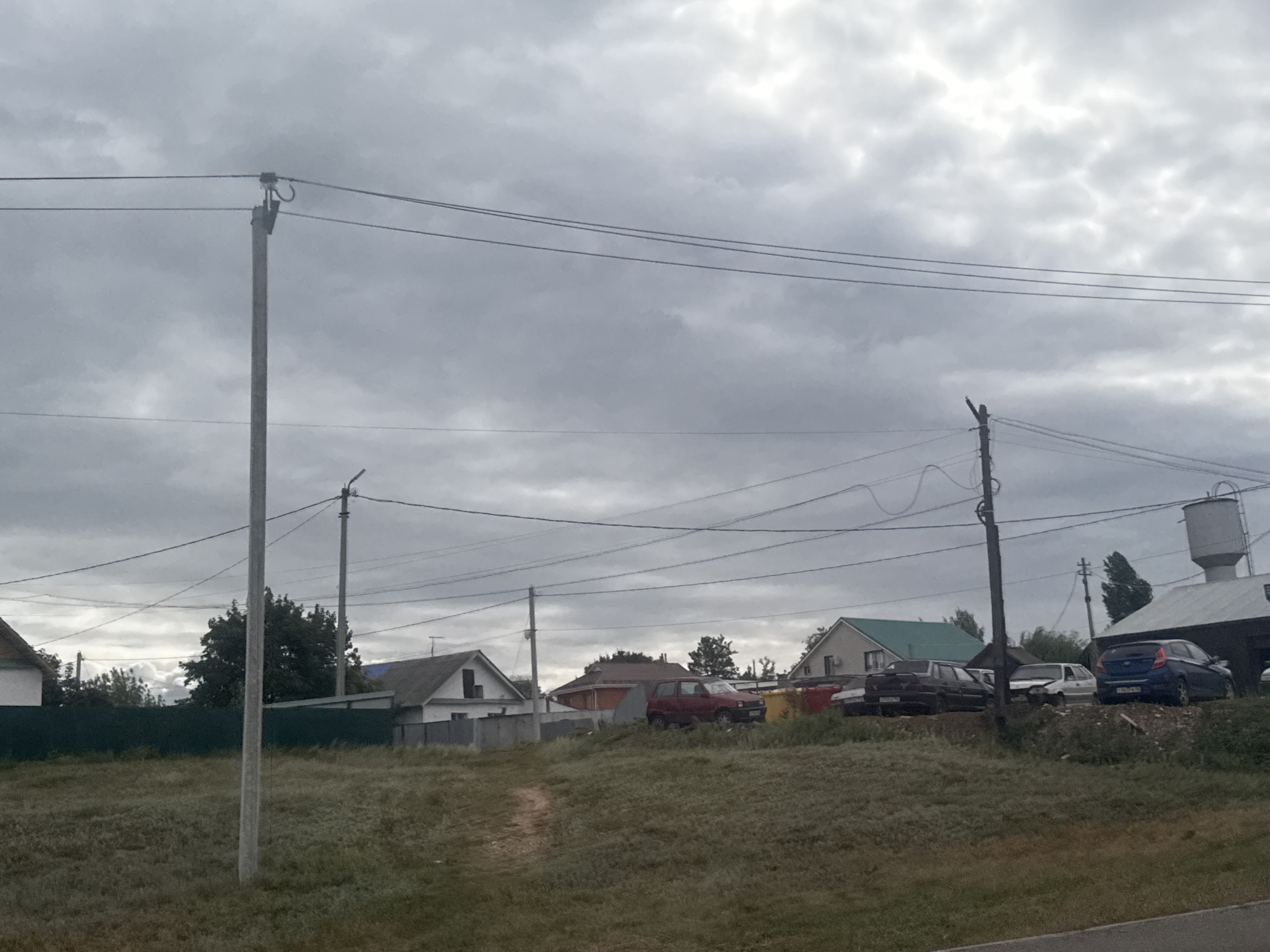 The image depicts a street scene in Novosergiyevka, Russia, under an overcast sky. The foreground is dominated by a grassy field with a dirt path winding through it. A tall, concrete utility pole stands prominently in the left-center of the frame, with utility wires stretching across the upper portion of the image.
In the background, several residential buildings are visible. To the left, a white house with a darker roof is partially obscured by a dark green fence. Further to the right, a red car is parked on a slight incline, with other vehicles, including a dark-colored sedan and a blue hatchback, also present. A tall, cylindrical water tower is visible on the right side of the image, adjacent to a dark-roofed building. Trees are scattered throughout the scene, adding to the rural character of the location. The overall atmosphere is subdued due to the cloudy weather. No people are visible in the image.