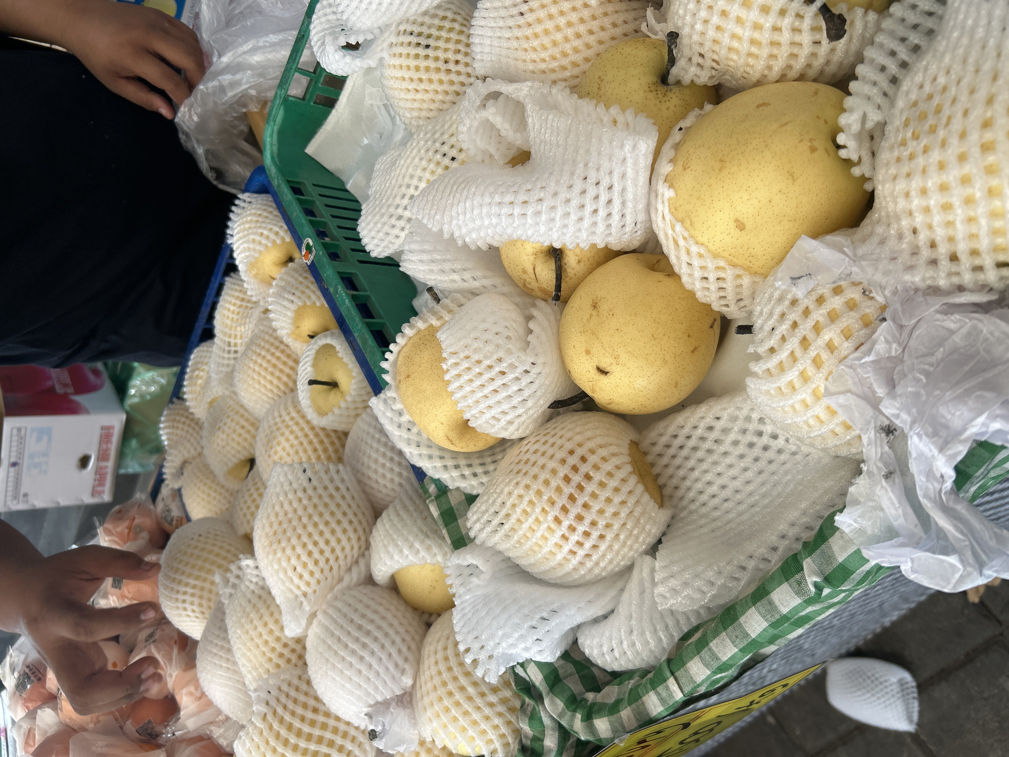 This image depicts a fruit stall, likely in Sukawati, Indonesia, showcasing a large quantity of pears. The pears are individually wrapped in white protective netting, a common practice for market fruit to prevent bruising. They are arranged in crates, one green plastic crate is visible at the top right, and another, green and white checkered fabric lined basket-like structure is seen in the foreground. Several pears are visible without netting, revealing their yellow skin and round, slightly elongated shape.
A person's hands, with darker skin tone, are seen reaching into the stall, presumably to select or arrange the fruit. The person is wearing a dark t-shirt. In the background, a portion of a cardboard box with some text, possibly indicating a brand or origin, is visible, along with other produce wrapped in plastic. The ground appears to be paved with square gray tiles. The overall scene suggests a bustling market environment where fresh produce is being sold.