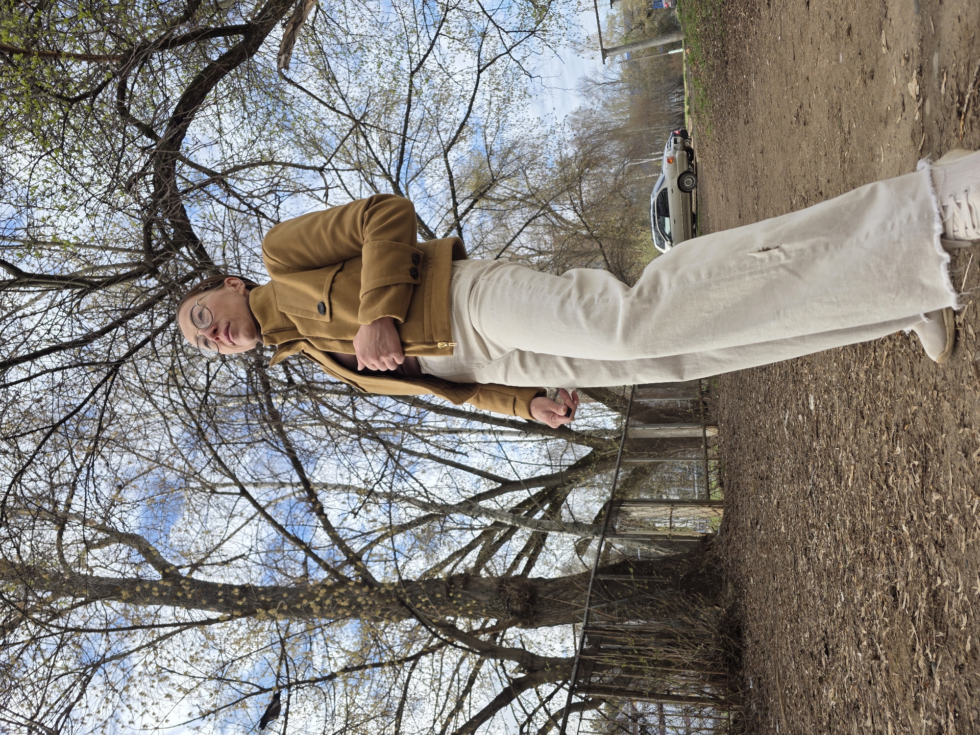 A full-length outdoor photograph shows a person standing on dirt. The person is wearing a brown jacket, beige pants, and beige shoes. The pants have a hole in the thigh. A tree with bare branches is behind the person. In the background, a car is parked on a dirt road. The location appears to be in Chaikovsky, Russia.