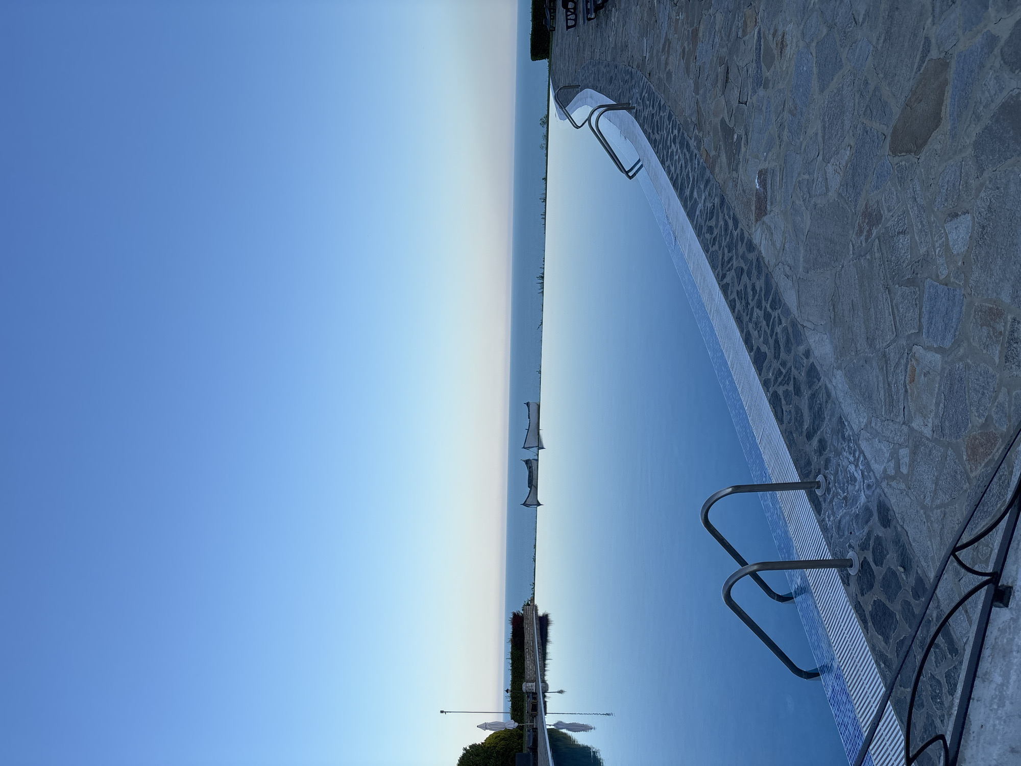 The image shows an infinity pool overlooking the Black Sea in Topola, Bulgaria. The sky is a clear, pale blue, transitioning to a lighter hue near the horizon. Two canoes are visible on the calm, reflective surface of the sea. To the left of the pool, there's a white umbrella, lounge chairs, and lush green bushes. The pool itself is a deep blue, with metal railings and steps leading into the water. The pool deck is paved with irregular, grey flagstones. The overall scene evokes a sense of tranquility and serenity.