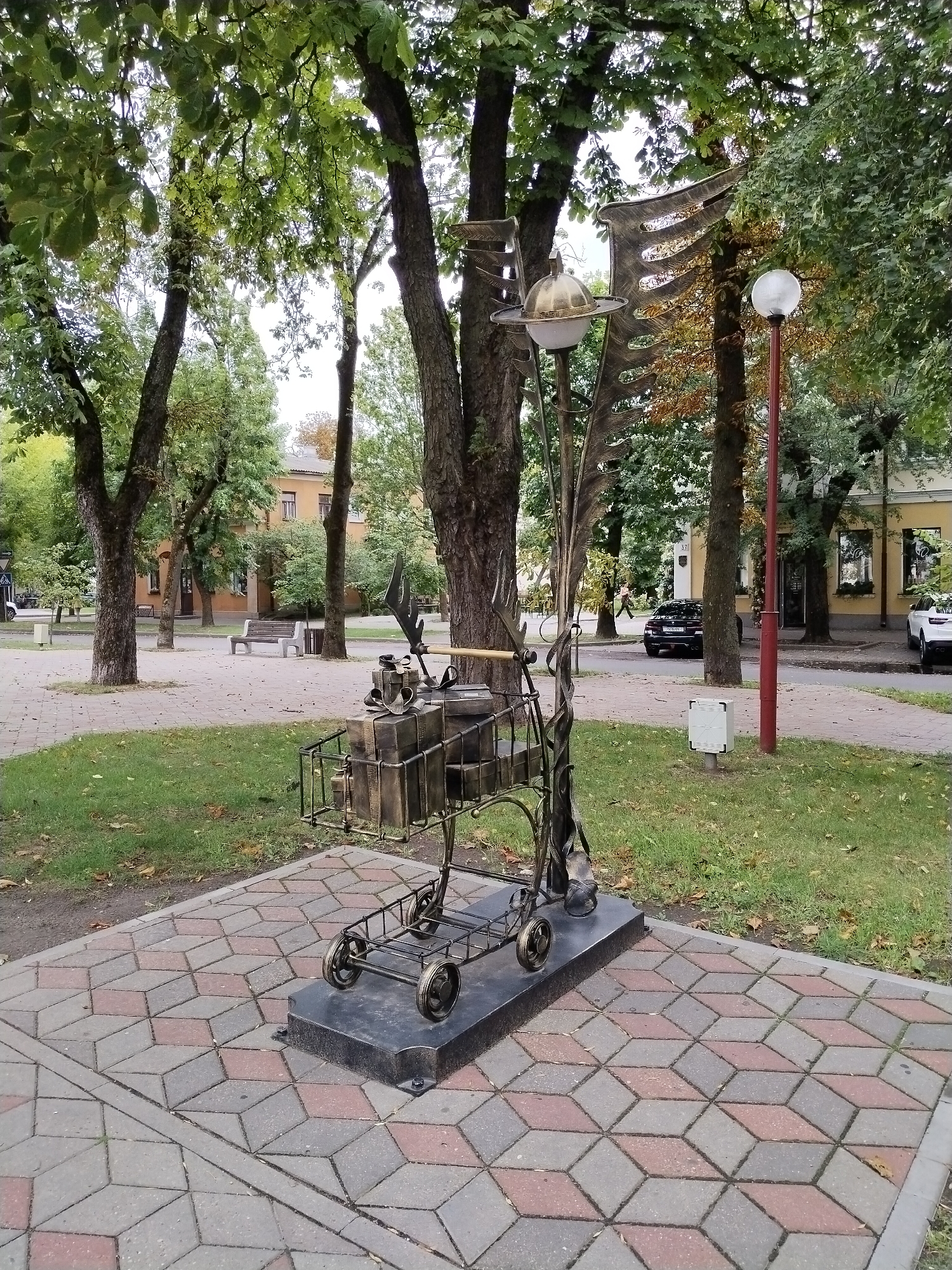 A metal sculpture resembling a shopping cart filled with various objects is positioned on a patterned paving stone area in an urban park setting. The sculpture features intricate metalwork, including decorative elements that extend upwards, resembling wings or a stylized tree. A sphere, possibly representing a planet or a light fixture, is attached to the upper part of the sculpture. Behind the sculpture, trees with green foliage are visible, some displaying autumn colors. A lamppost with a spherical light cover stands to the right of the sculpture. In the background, there are buildings, a street with cars, and park benches. The paving stones are arranged in a geometric pattern of rhombuses and squares in shades of gray and red. This sculpture is located in Brest, Belarus, and is a part of the city's public art installations.