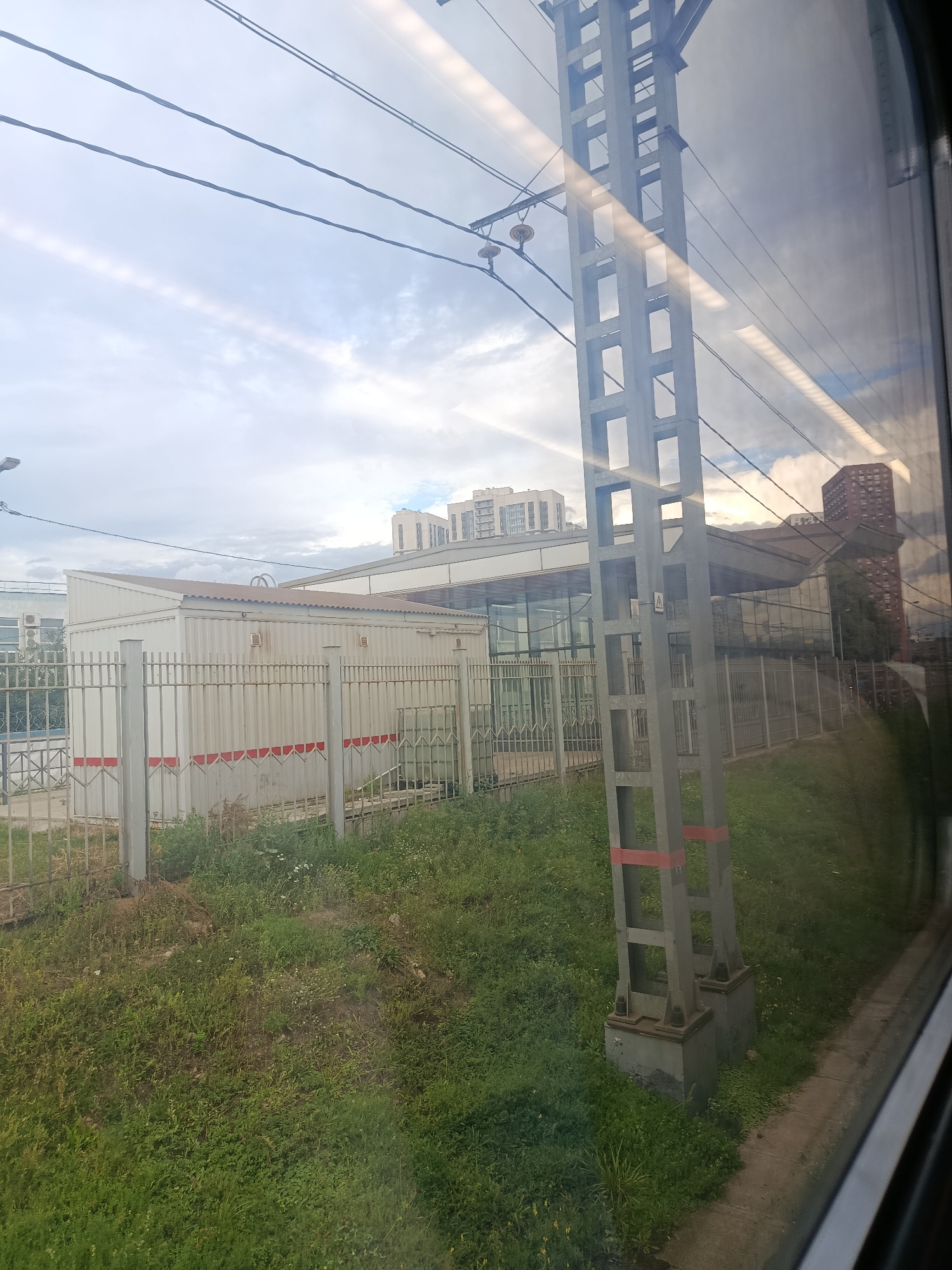 The image captures a view from inside a train, looking out at an urban landscape under a cloudy sky. In the foreground, a tall, grey metal pylon with rectangular cutouts supports overhead electrical wires, a common sight along railway lines. The pylon is marked with horizontal red stripes near its base. To the left of the pylon, a white, corrugated metal building with a sloped roof is visible behind a rusty metal fence. The building has several ventilation grates and is adorned with horizontal red stripes. Beyond the building, a modern structure with a glass facade and a dark roof is partially obscured. In the distance, several multi-story residential buildings, some appearing to be of modern construction with light-colored facades, are visible. A tall, reddish-brown building with multiple windows stands to the right of the glass-fronted structure. The ground is covered in green grass and various low-lying plants. The overall impression is of an industrial or semi-urban environment adjacent to a railway line, likely within Moscow, Russia, given the characteristic architecture and infrastructure.