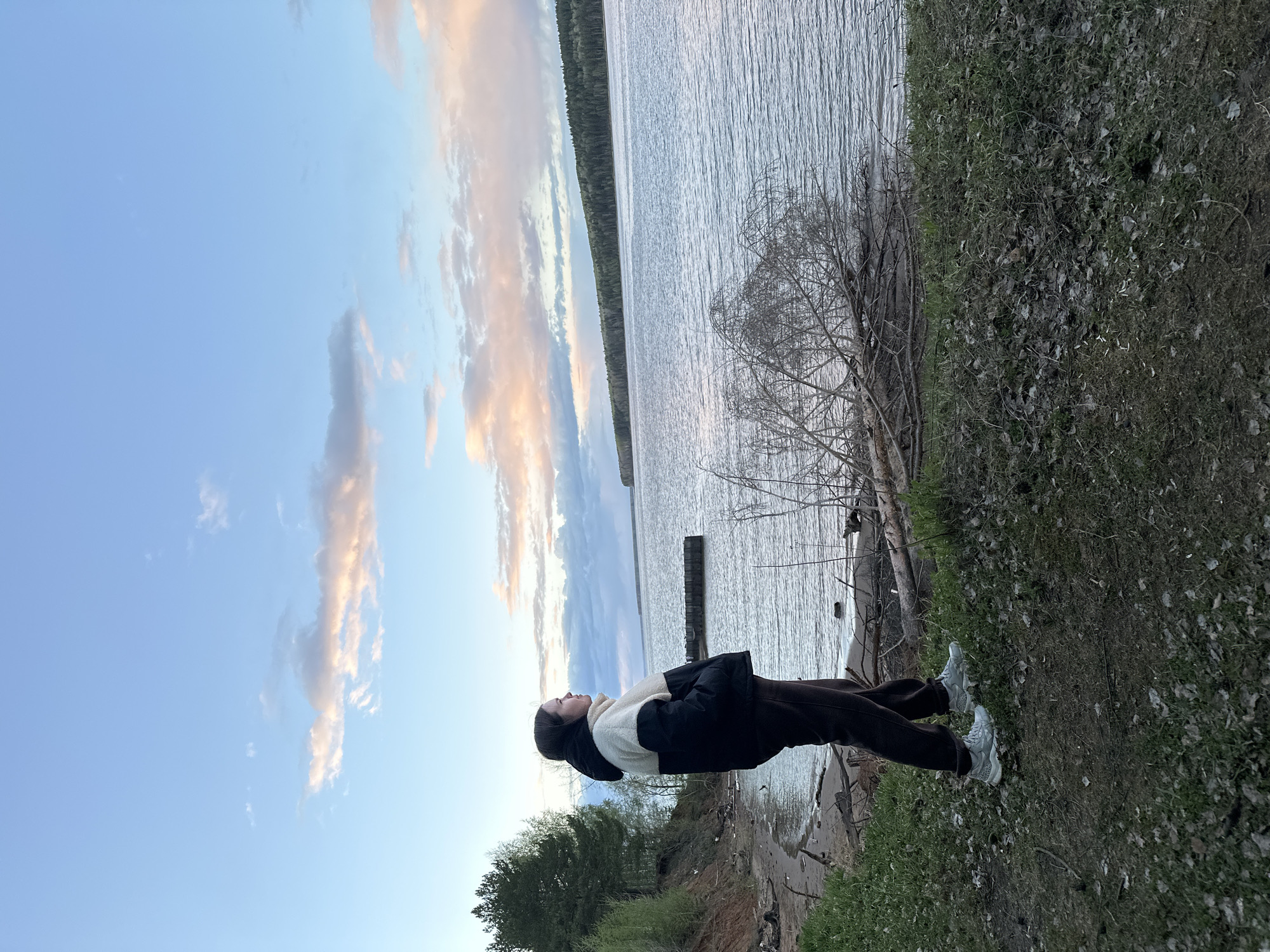 A vertically oriented medium shot captures a person standing on a grassy embankment, looking upwards towards a cloudy sky. The person is wearing dark pants, a black jacket with a light-colored interior, and light-colored shoes. They appear to be in a contemplative pose, gazing at the sky.
Behind the person is a body of water, possibly a lake or a wide river. A strip of forest is visible on the opposite bank, under the sky. The sky is partially covered with clouds that are illuminated with warm tones, indicating either sunrise or sunset.
The embankment is covered with grass and some bare patches, with a few bare tree branches visible. The location is Chaikovsky, Russia.