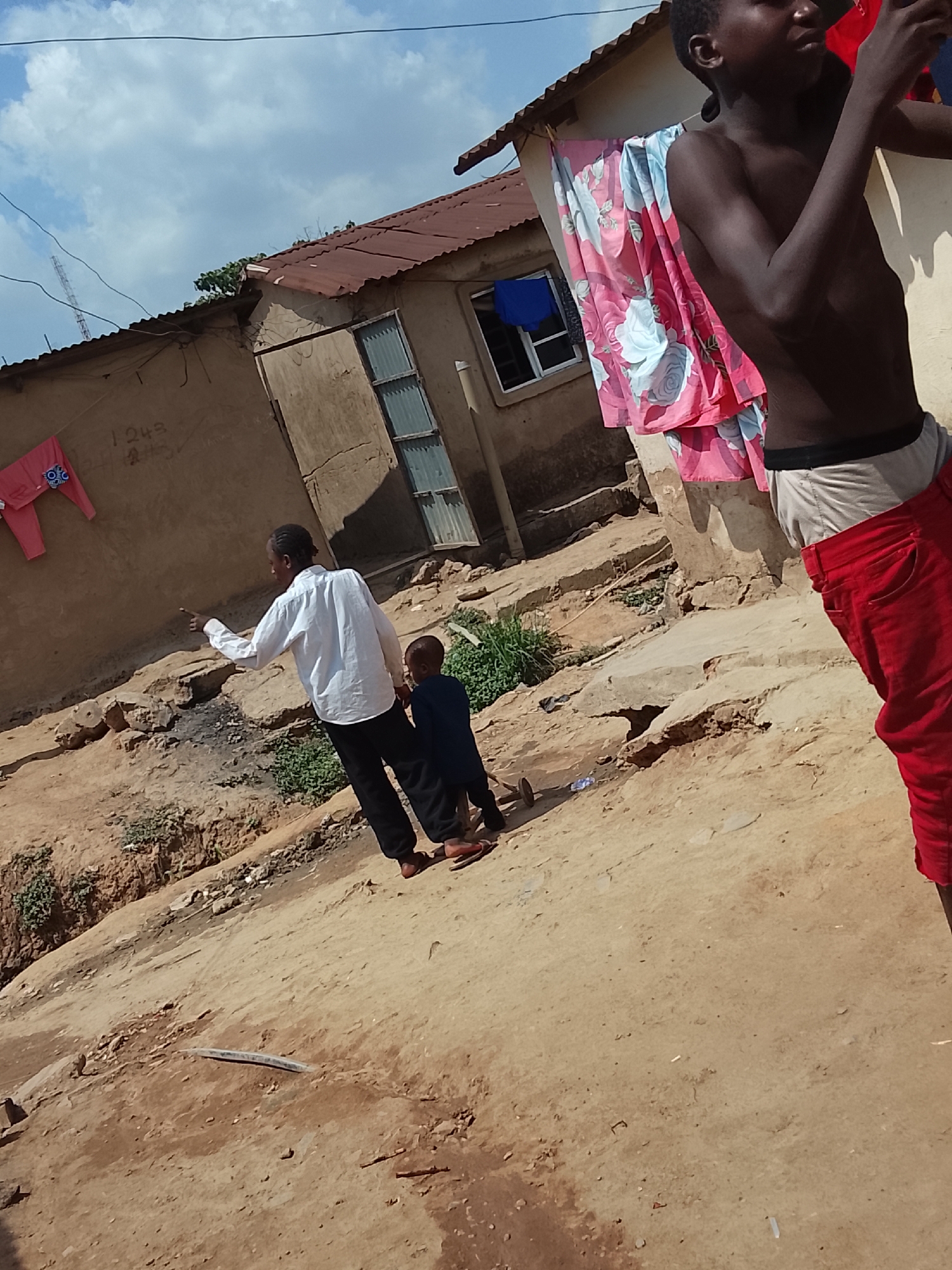 The image shows a dirt street in what appears to be a low-income area in Abuja, Nigeria. A boy in red pants and no shirt stands in the foreground to the right, partially obstructing the view. Behind him, hanging outside a building is a patterned pink cloth. Further back, a person in a white shirt and black pants points towards the building next door, accompanied by a small child wearing dark clothing. The buildings are constructed of mud or concrete, with corrugated metal roofs. Laundry hangs on the side of one building. The ground is dirt, and there is some sparse vegetation.