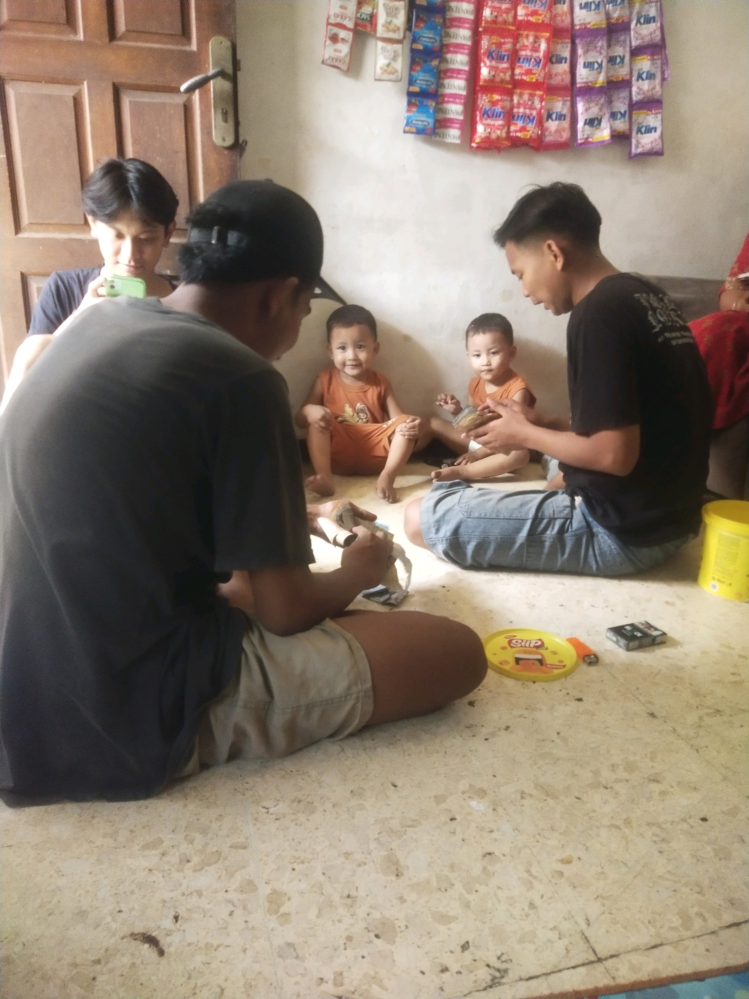In Surabaya, Indonesia, a group of individuals, including two children, are gathered in a room with a tiled floor and a wooden door. Two young boys, one wearing an orange shirt, sit on the floor. Two young men are also present; one is on his phone, and another, wearing a black shirt and denim shorts, is holding a small item in his hands. The room has a plain wall adorned with colorful sachets of products, possibly hygiene or food items, suggesting a domestic or small shop environment. On the floor, various objects are scattered, including a yellow plate with text, a cigarette lighter, and a pack of cigarettes. A large yellow bucket is visible in the background, near the wall. The lighting appears to be natural, coming from an unseen source, casting soft shadows. The overall scene depicts a casual gathering of people in a relatively simple interior setting in Surabaya.