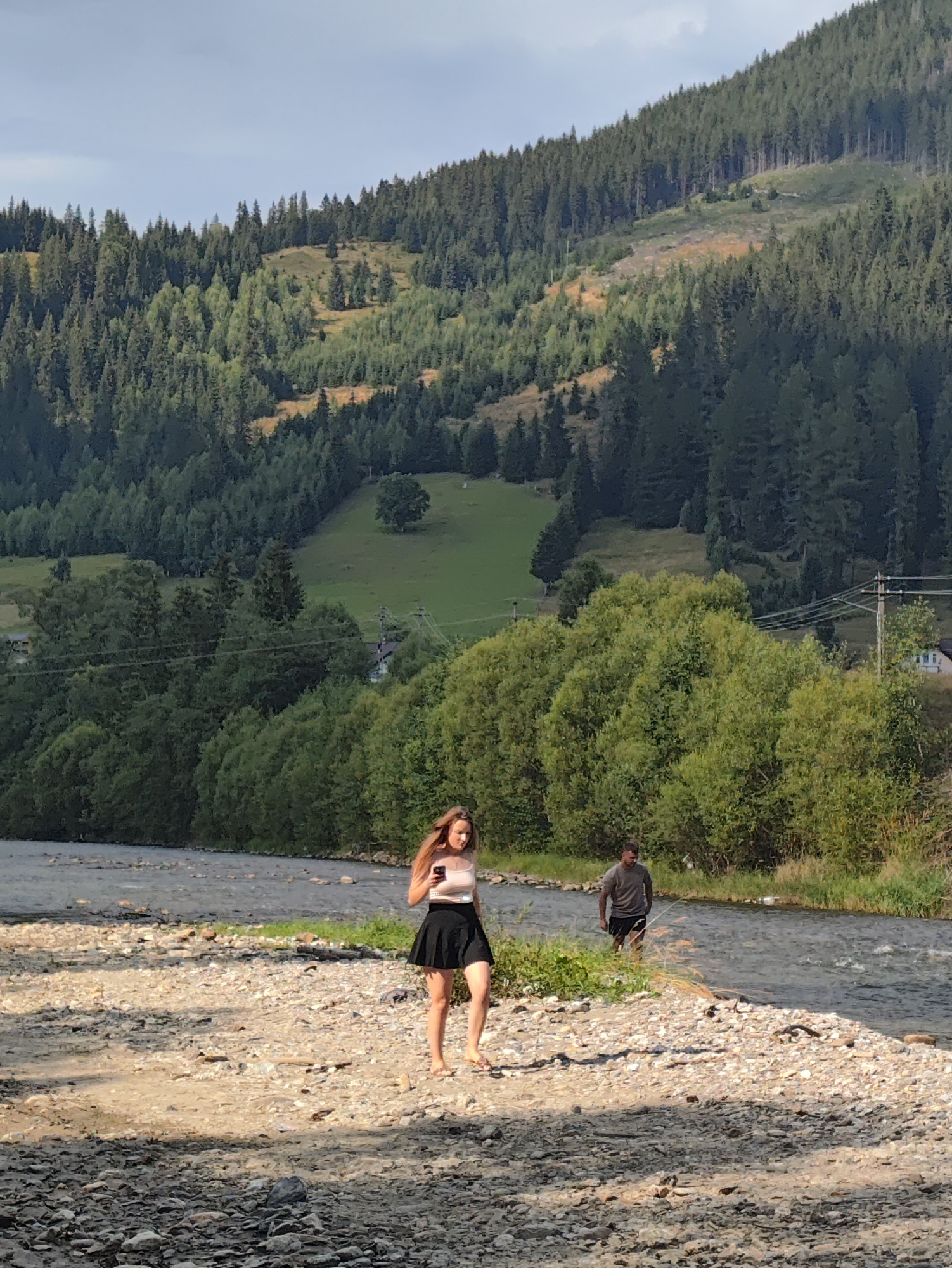 A young woman with long brown hair, wearing a white tank top and a black skirt, walks on a rocky riverbed, looking at her phone. To her right, a man in a grey t-shirt and black shorts stands in the shallow river. The river flows past a gravelly shore and is bordered by lush green trees. In the background, a steep, tree-covered mountain rises under a cloudy sky. The scene appears to be in a natural, outdoor setting, possibly near Botoș, Romania, given the resemblance to the Carpathian foothills, known for their mountainous terrain and dense forests.