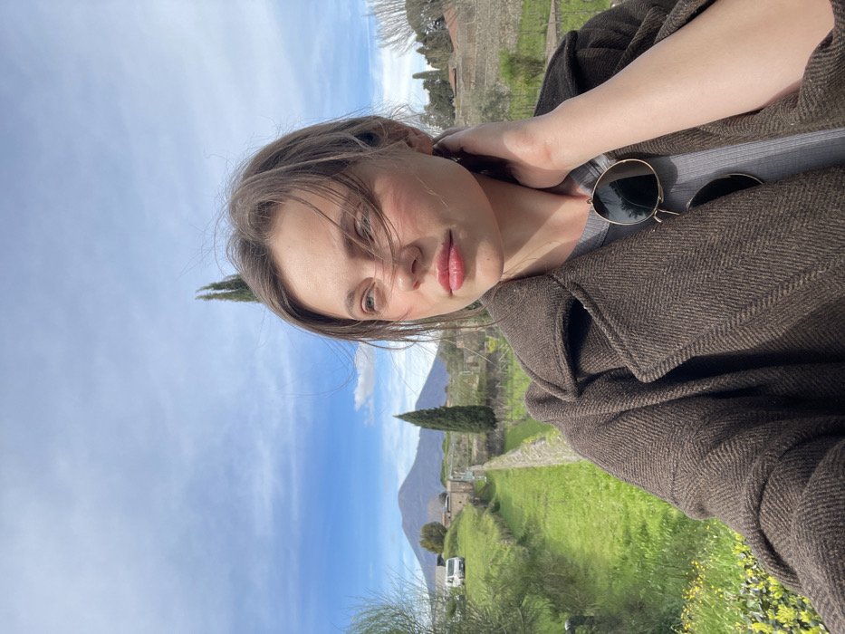 A young adult female is positioned in the foreground of an outdoor scene within the archaeological site of Pompeii, Italy. She has light brown hair, with strands disturbed by wind, and exhibits a light complexion with subtle facial flushing. She wears a dark brown herringbone-patterned wool coat over a grey ribbed top. Dark sunglasses with gold frames are suspended from her neckline. Her right hand is placed on the back of her neck. Her gaze is directed off-camera to the viewer's upper left.
The background features a distinct, long, green pathway or vineyard row, bordered by a low stone wall on its right side, receding into the distance. Multiple tall, slender cypress trees are visible along this pathway. In the mid-ground, characteristic ancient Roman ruins are discernible, and a white van is parked on a grassy area further down the path to the left. The prominent, conical peak of Mount Vesuvius dominates the distant horizon under a bright blue sky with scattered white clouds.