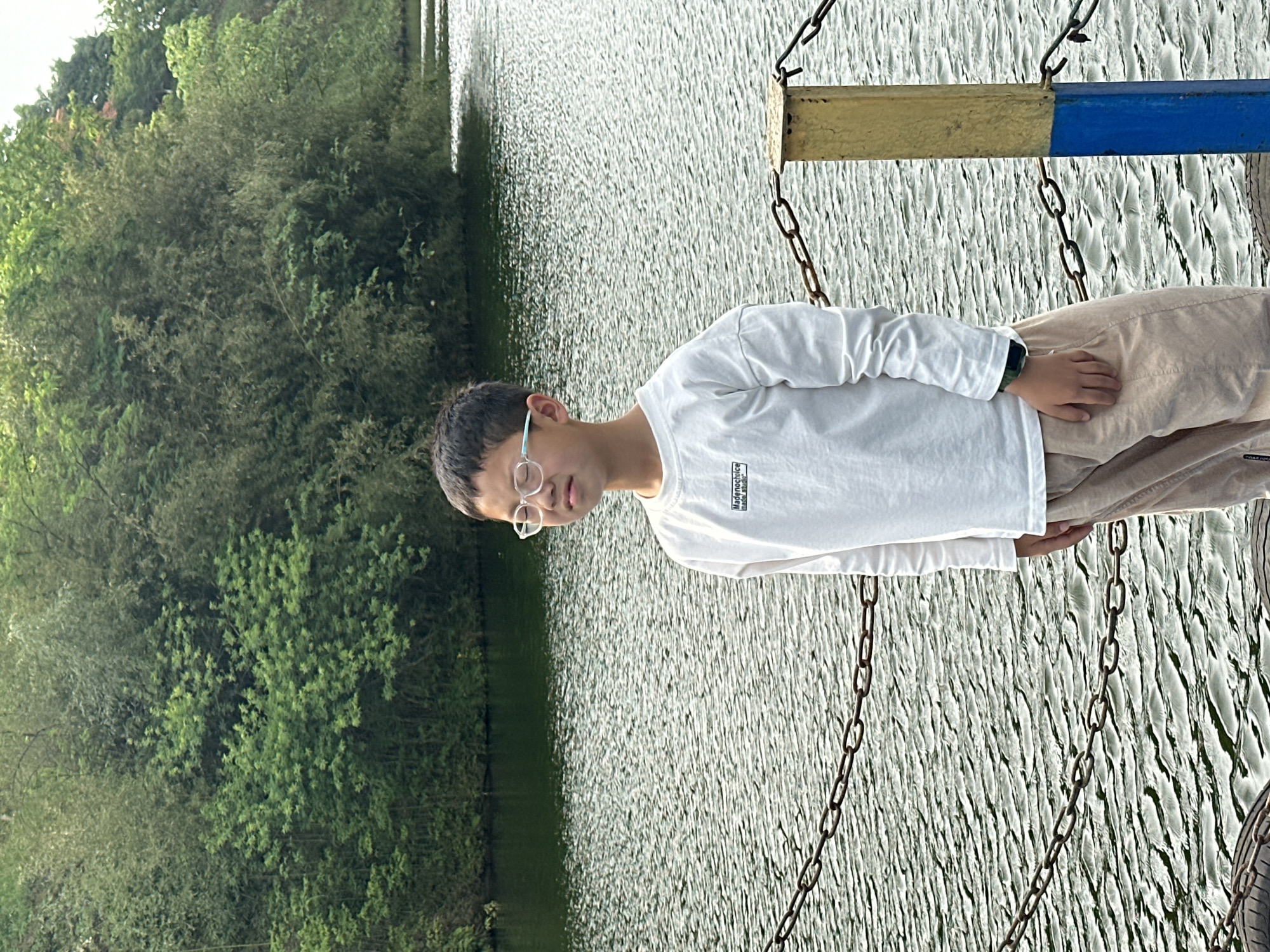 A person wearing a white long-sleeve shirt and light brown pants stands on a structure with metal chains and a yellow and blue beam. Behind the person is a body of water with trees on the opposite bank. The scene is likely in Liangjiang, China, given the prompt's indication.