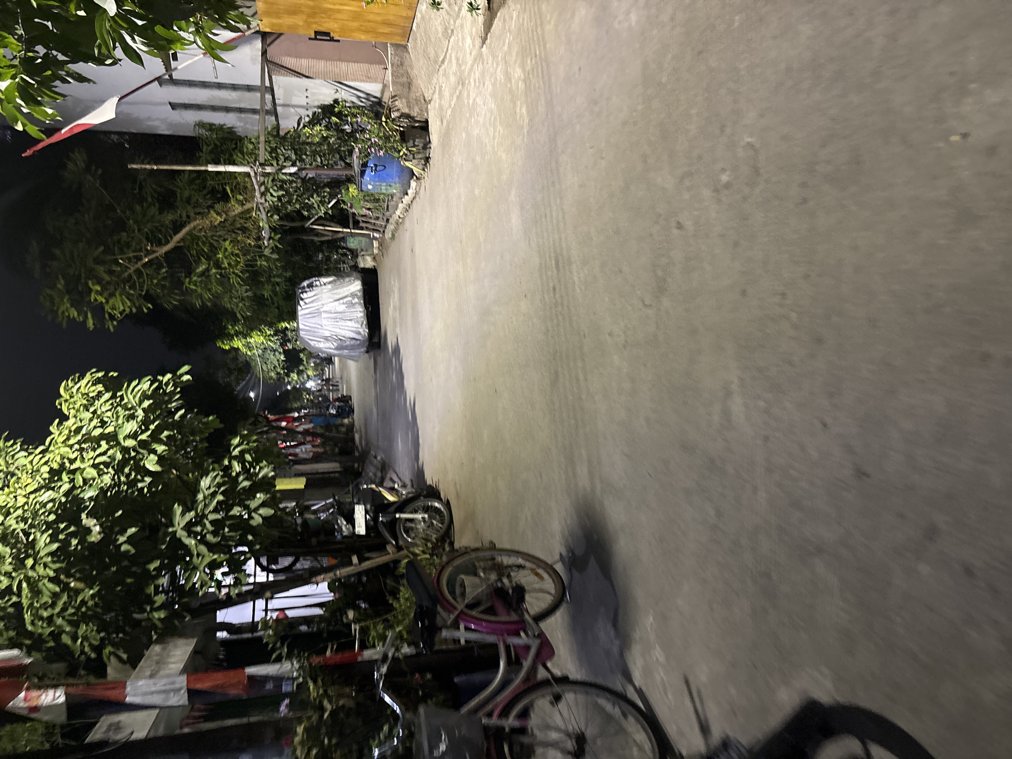 In Telagamurni, Indonesia, a street scene unfolds at night. The foreground is dominated by a pink bicycle parked on the left, with its front wheel partially visible. Further down the street, a silver-colored car is covered with a tarp. On the right side of the street, a building with a light-colored facade stands, and a red and white Indonesian flag is prominently displayed. Various trees line the street, their leaves illuminated by unseen light sources. Bicycles and motorcycles are parked along the roadside. A blue drum and some potted plants are also visible near the building on the right. The street surface is paved, with some irregularities and shadows indicating the time of day and the lighting conditions. The overall impression is of a quiet residential street in the evening.