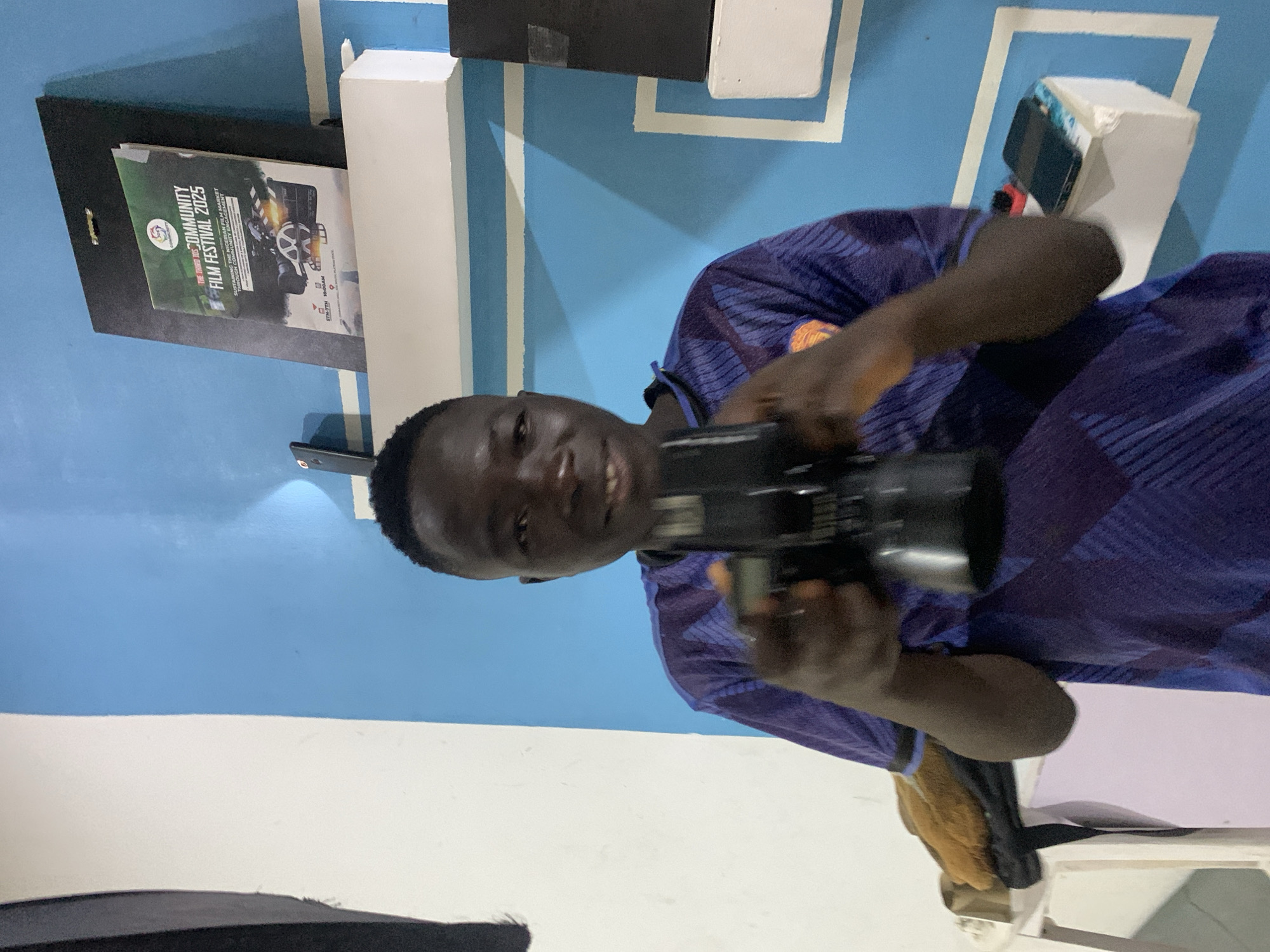 A smiling man with dark skin holds a black camera in his hands. He's wearing a blue and purple patterned shirt. Behind him is a blue and white wall. On the wall is a poster for the "The Third 10S Community Film Festival 2025," which is happening from the 5th to the 7th. The text on the poster reads "Sustaining the Nigerian Film Market". The location may be Gura Top, Nigeria.