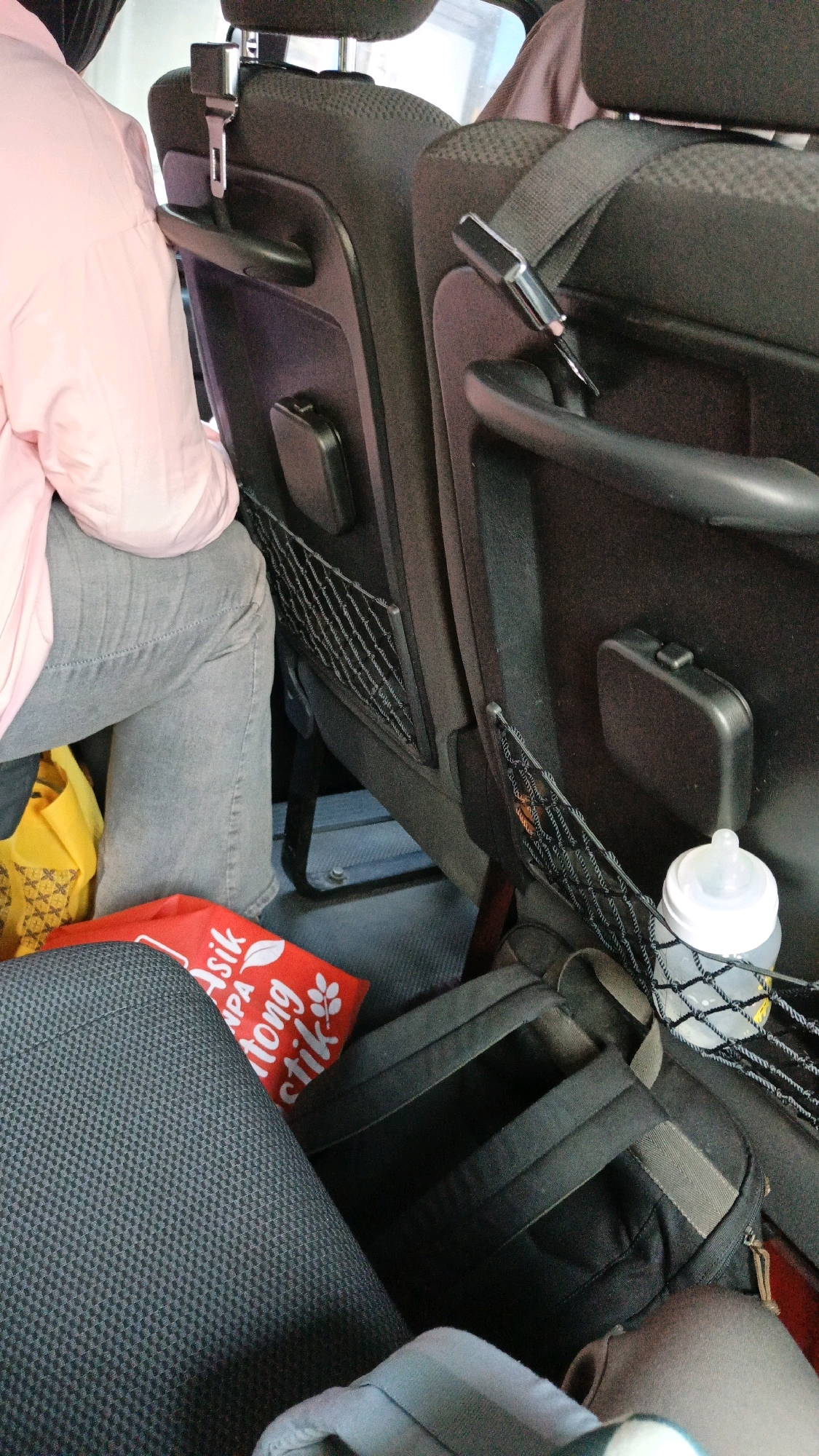 The image shows the interior of a vehicle, likely a bus or minivan, with a focus on the back seats. A person wearing a pink long-sleeved shirt and grey pants is seated on the left. In front of this person, a yellow bag with a patterned design is visible. Below the seats, a red bag with white text and floral designs is placed. To the right, a black backpack is situated, and a baby bottle with a white lid is tucked into a mesh pocket on the seatback. Seatbelts are visible, and the overall environment appears to be inside a mode of transportation, possibly en route within or near Weru Lor, Indonesia, given the context.