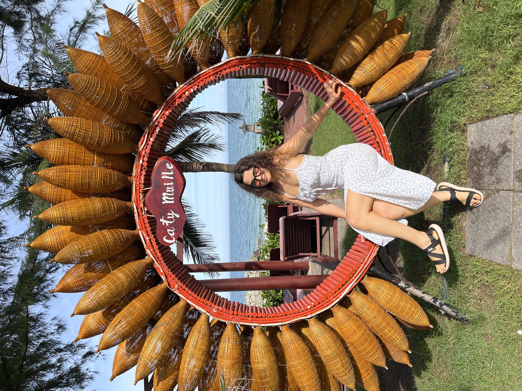 A woman with curly brown hair and glasses is sitting on a large, circular, sunflower-shaped art installation made of woven reeds and red bamboo. She is wearing a light blue and white floral print sundress and black sandals. The art installation has a sign that reads "Catty Mui Ne". In the background, a calm ocean is visible, with beach chairs and palm trees. The scene appears to be in Mũi Né, Vietnam.