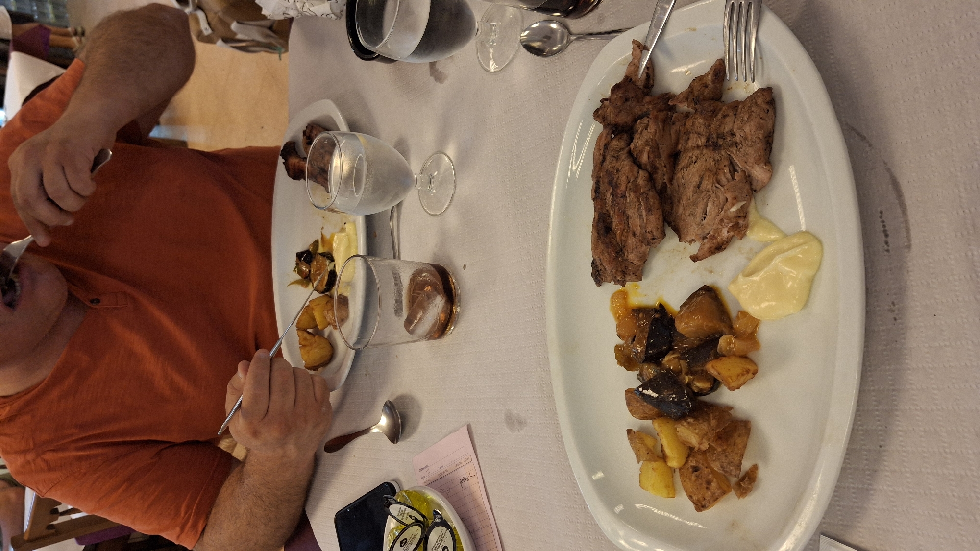 The image shows a close-up of a table setting at a restaurant in La Puebla de Híjar, Spain. A person, partially visible from the chest up, is wearing an orange t-shirt and eating from a plate of food. The plate contains sliced meat, roasted potatoes, and what appears to be eggplant. There are also glasses of water and ice on the table, along with cutlery and a small white bowl containing packets. The table is covered with a white tablecloth. The background suggests an indoor dining environment with other tables and chairs.