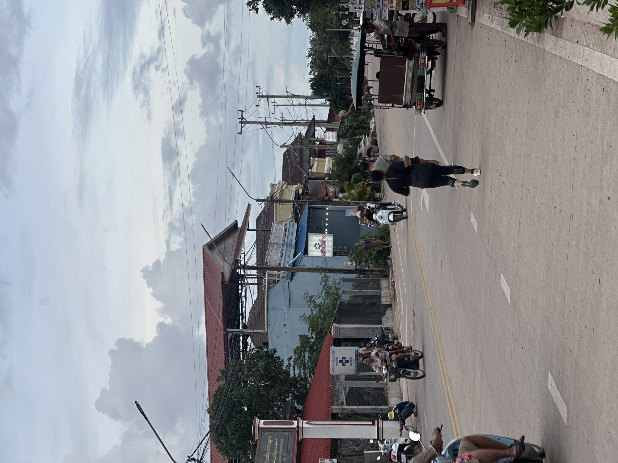 The image depicts a street scene in Kampot, Cambodia, under a cloudy sky. A paved road runs through the center, with a sidewalk on the right. Several vehicles are present, including motorcycles, a tuk-tuk, and a small blue motorbike. On the left side of the road, there are buildings with red and blue roofs, and a signpost. A person in a black shirt and black leggings is jogging on the right side of the road. Another person, wearing a light-colored t-shirt, is jogging behind them. A man in a light-colored shirt is standing near the tuk-tuk on the right side of the road. A person wearing a yellow hat is crouching near a parked white motorcycle on the left side of the road. The overall atmosphere is that of a typical street in a Southeast Asian town.