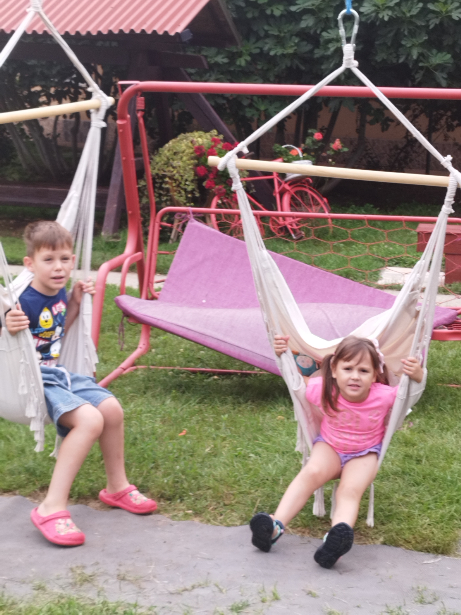 Two children are on swings in a backyard, with a red swing set and a pink hammock-like cushion visible in the background. A boy, wearing a blue t-shirt with cartoon characters and denim shorts, sits in a white hammock swing on the left. A girl, wearing a pink t-shirt and purple shorts, is in a similar white hammock swing on the right. They are both looking towards the camera. The environment appears to be a residential backyard with green grass and some foliage. The image quality suggests it was taken with a mobile phone camera.